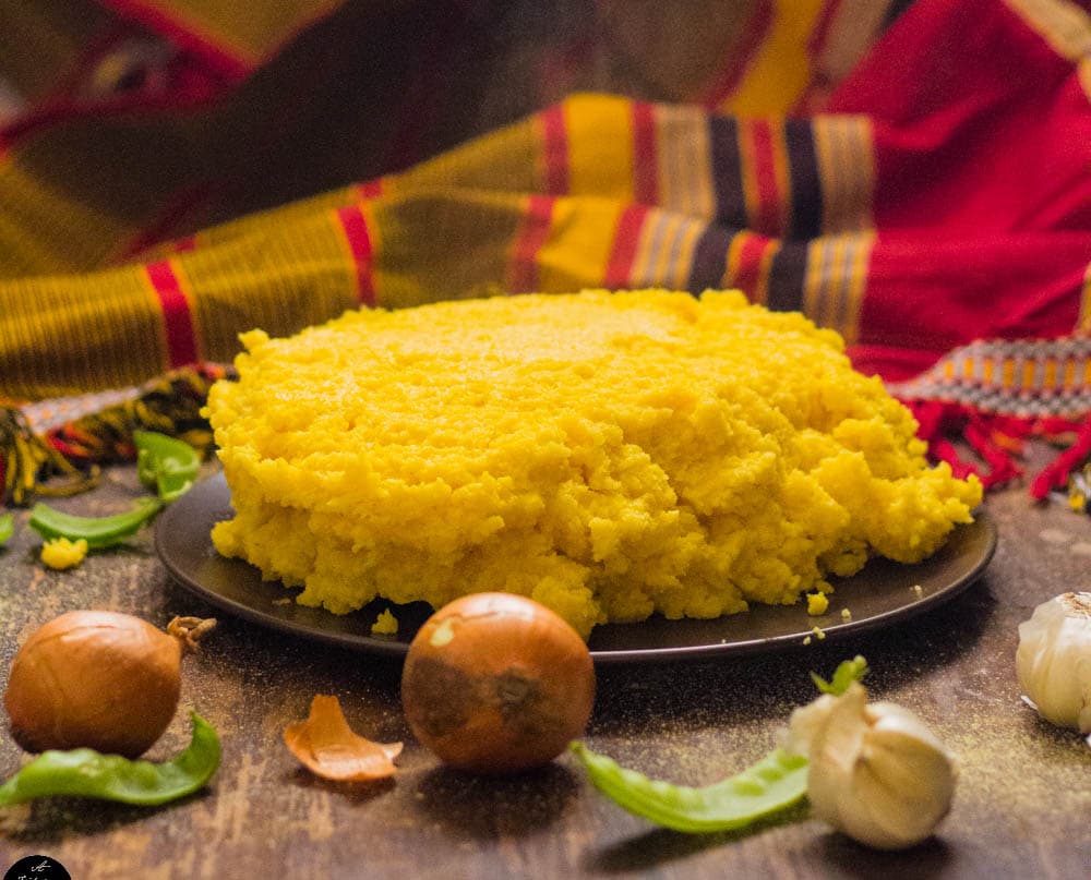 Mound of cooked yellow posho maize meal served on a plate with a woven cloth backdrop.
