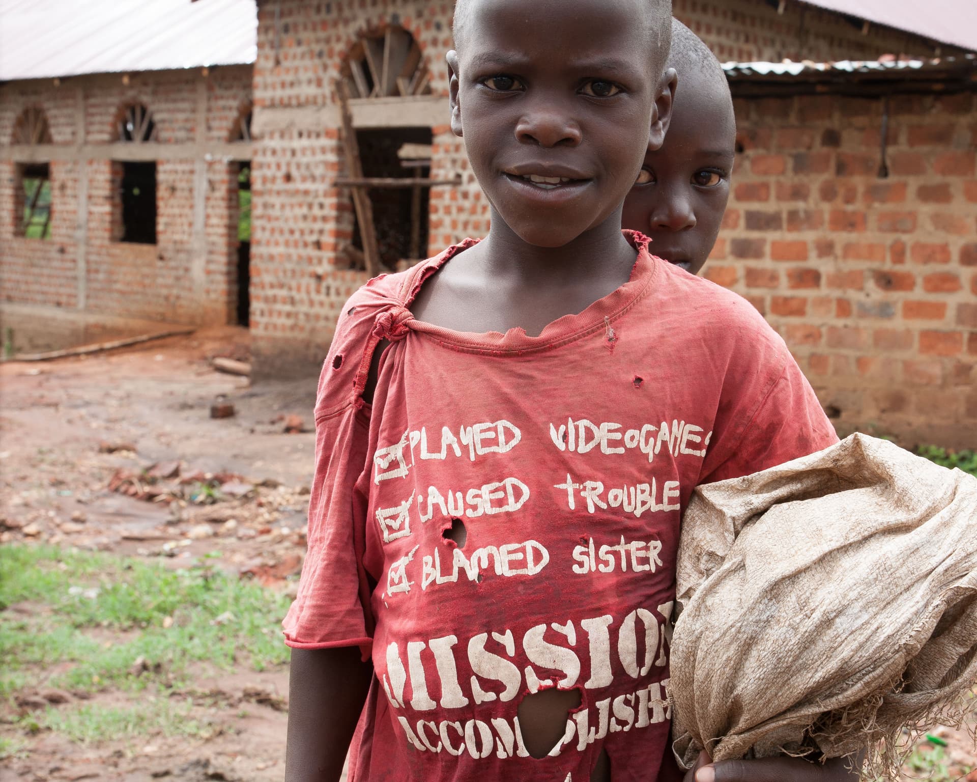Two boys stand in front of a brick building with graffiti-style writing, one wearing a red shirt reading Mission.