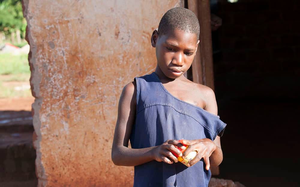 A young girl stands against a mud wall holding a small piece of food in her hands.