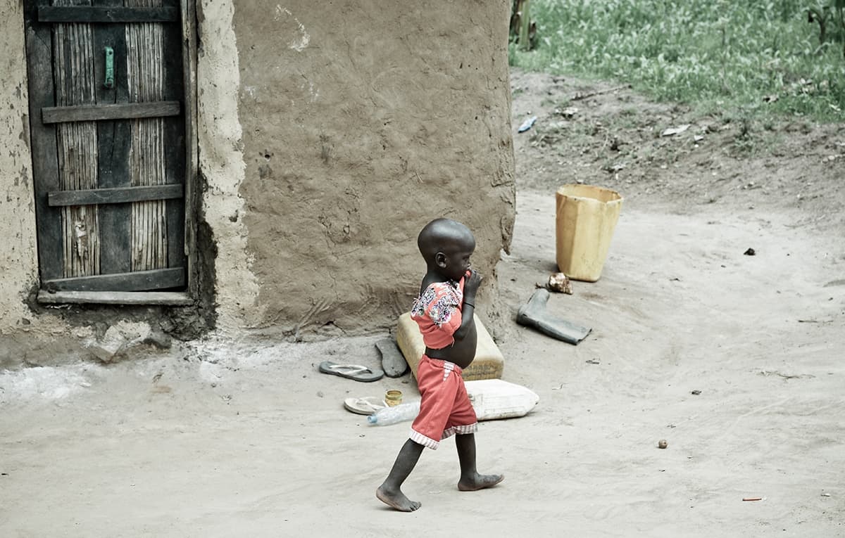 A child in a red dress walks outside near a mud building and yellow jerrycans.