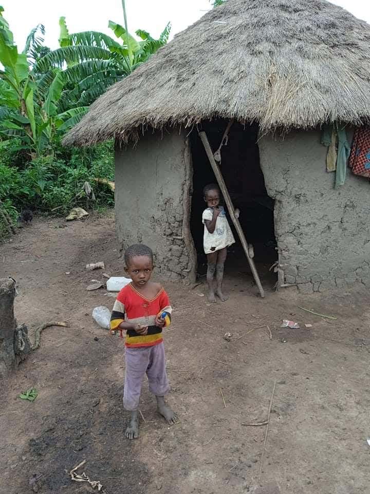 A child stands outside a thatched mud hut while another child looks out from the doorway.