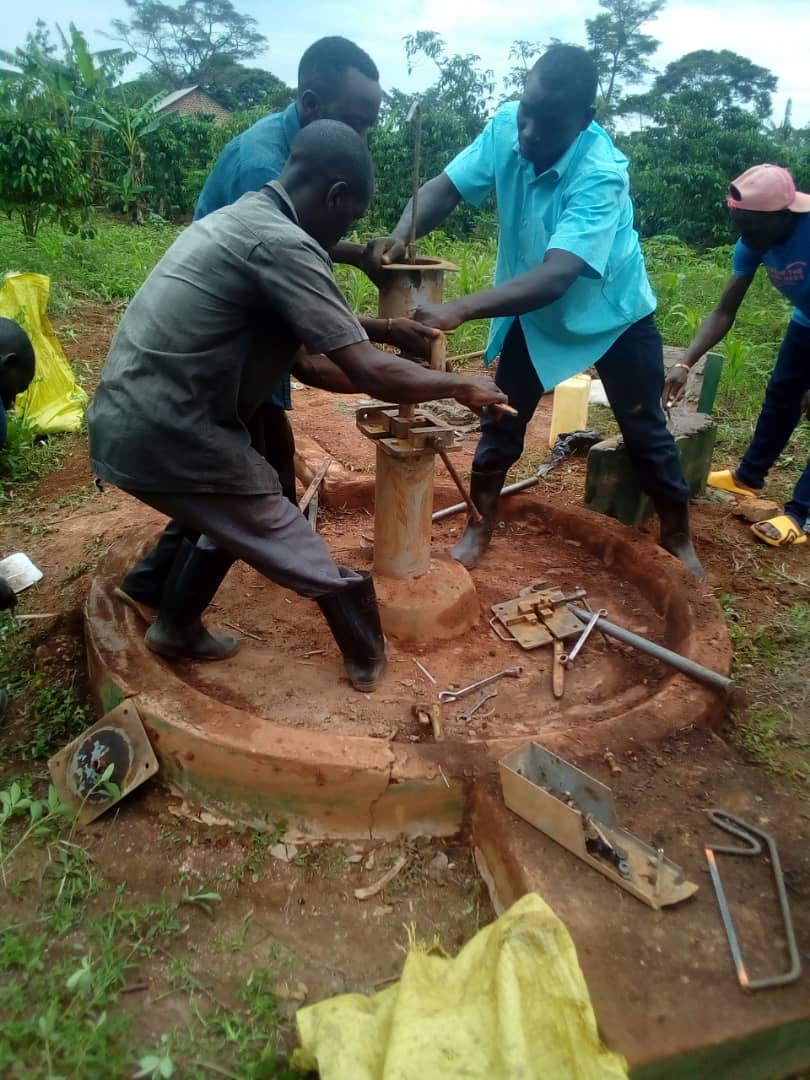 Two men use tools to repair a hand-pump water well, with onlookers and yellow jerrycans nearby.