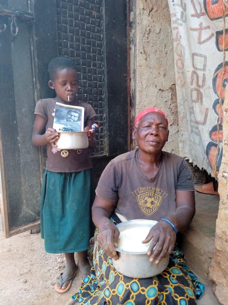 A girl holds a bowl of rice while an elderly woman crouches preparing food nearby.