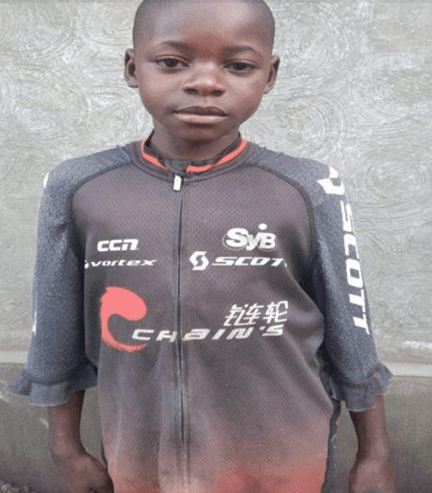 Portrait of a boy in a gray zip-up jacket with graphic branding standing against a wall.