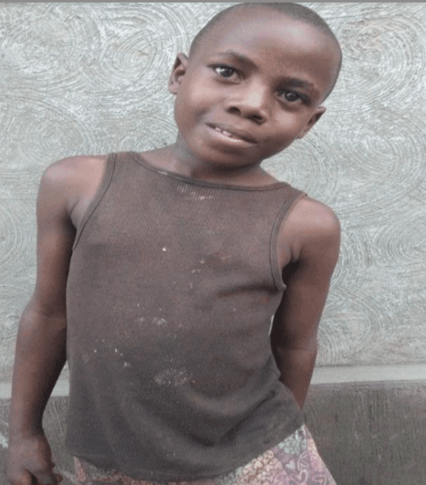 Portrait of a boy in a worn tank top standing against a concrete wall.