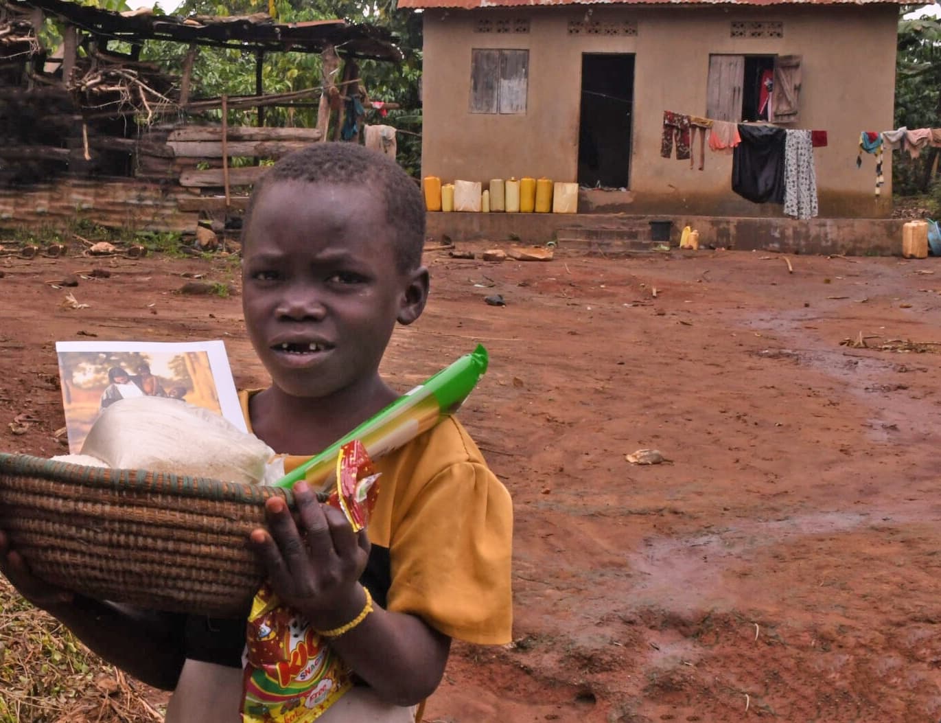 Boy standing outside a mud-brick house holding a woven basket with a JFM food gift package.