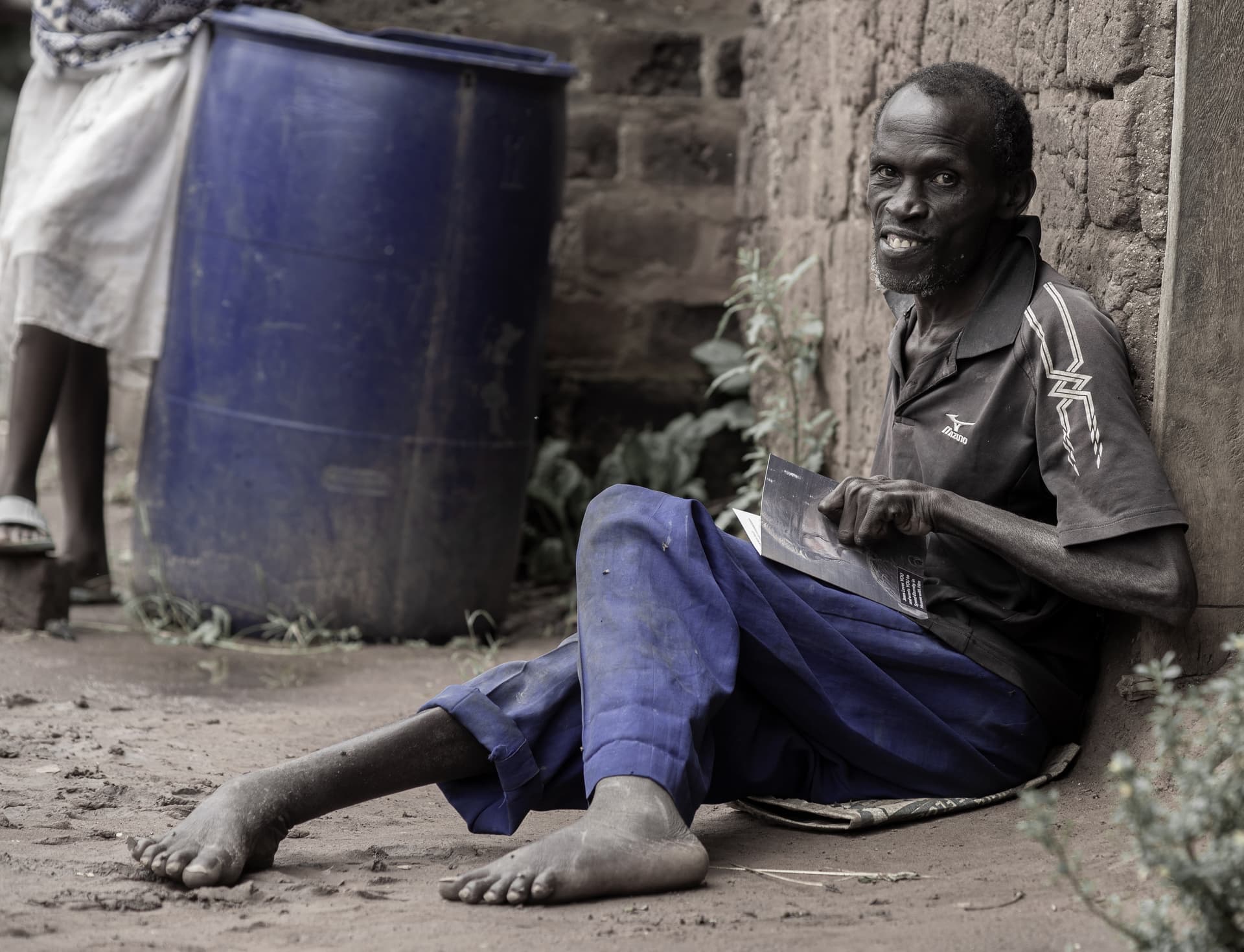 Black-and-white photo of a man, likely Kalindi Moses, lying on the ground near blue plastic barrels.