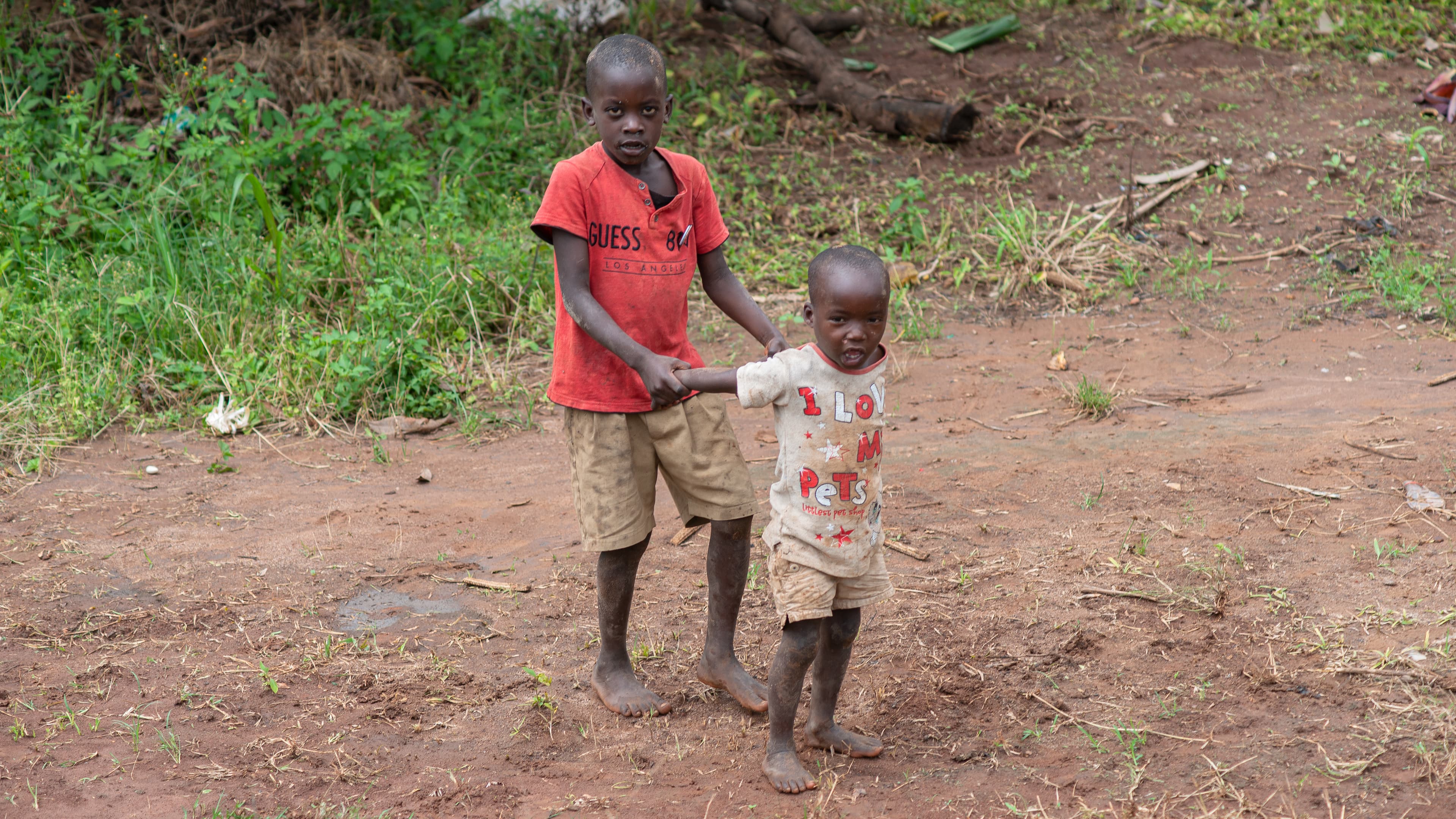 Two children stand together on a dirt path, one in a red shirt.