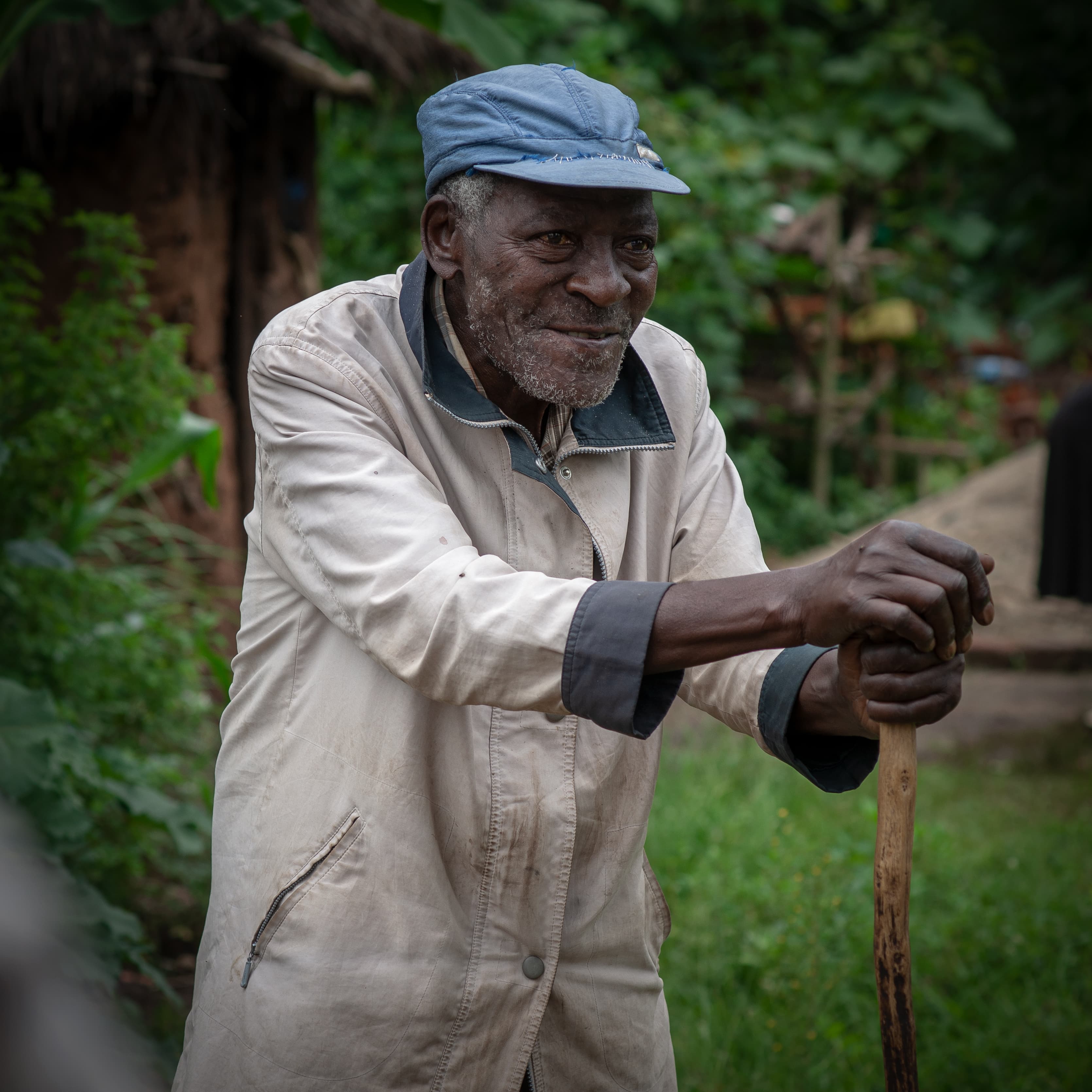 An elderly man leaning on a cane stands near a mud-brick building.