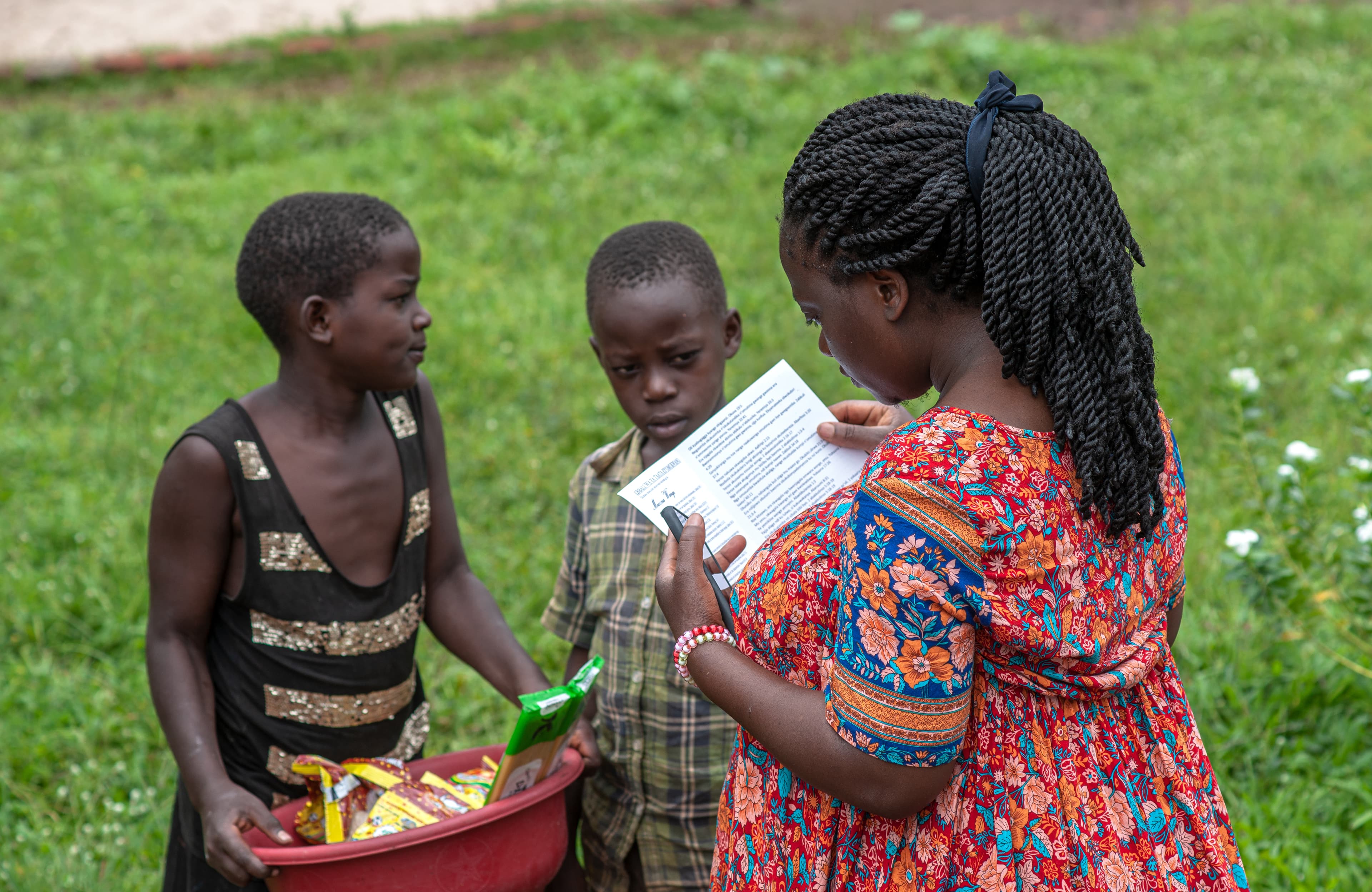 Three children stand together outdoors, one holding a red bowl of food.