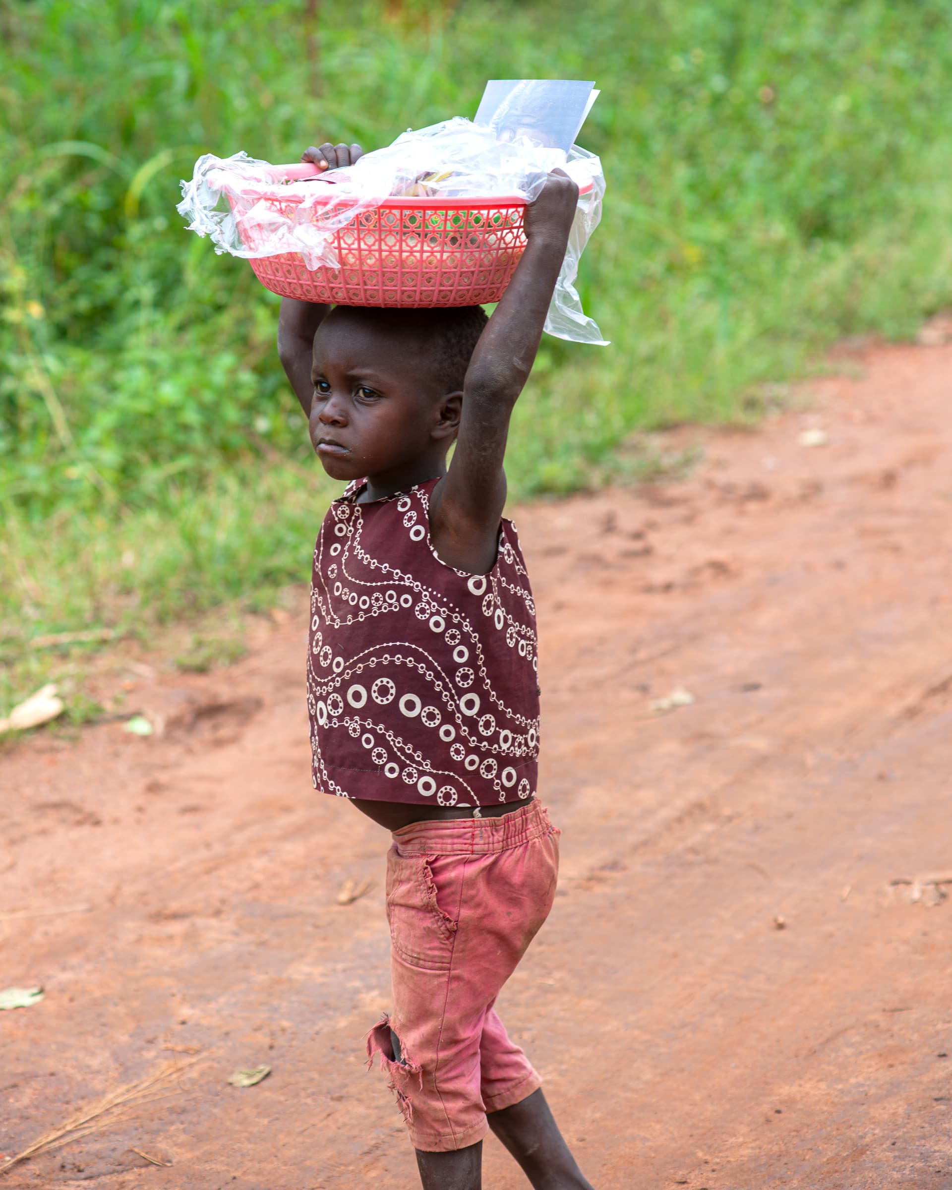 A girl carries a white bundle on her head along a dirt road.