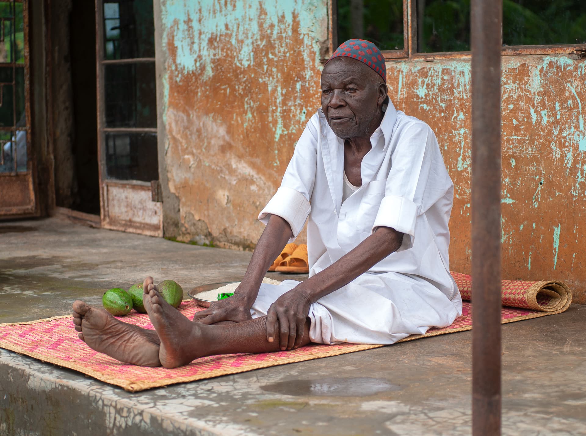 An elderly man in white sits on a porch beside a bundle of belongings.