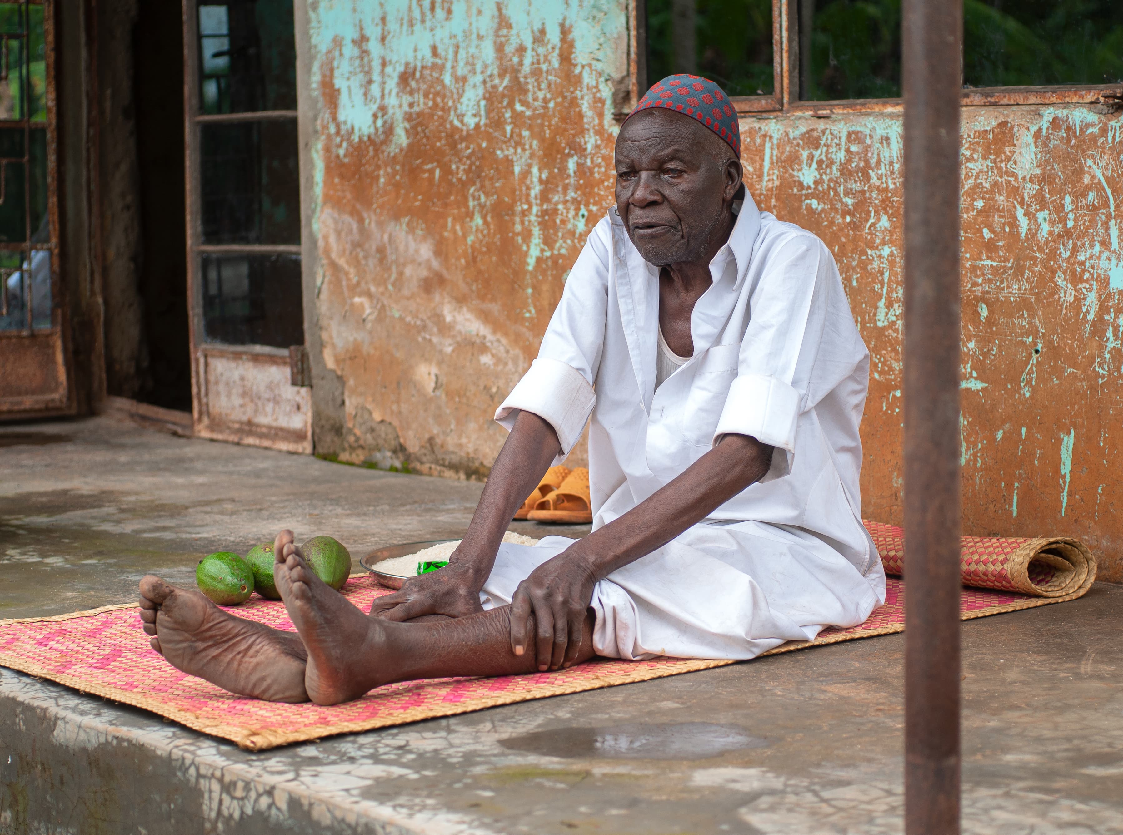 An elderly man in white sits on a porch beside a bundle of belongings.