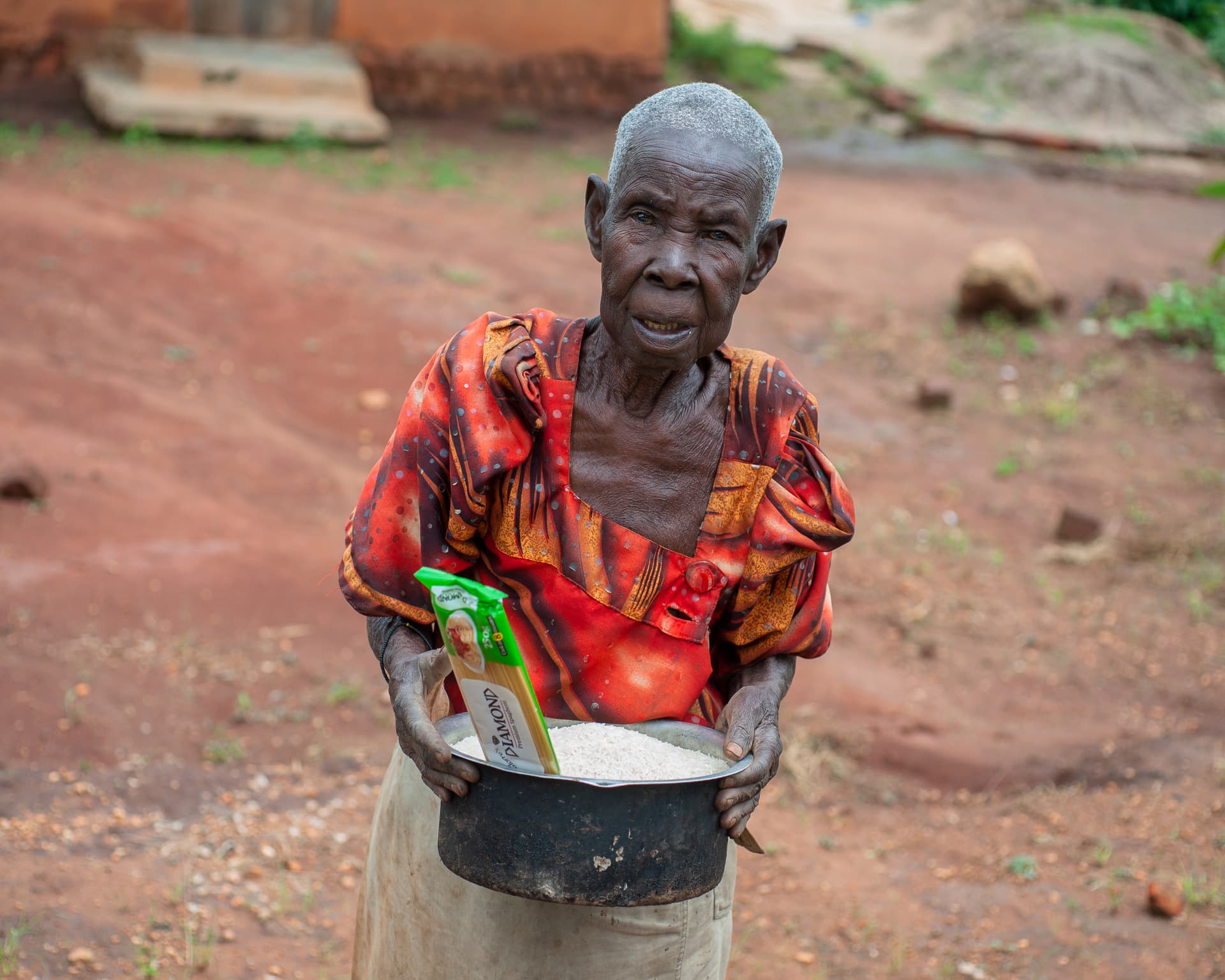An elderly woman in a patterned top holds a bowl with a photo card inside.
