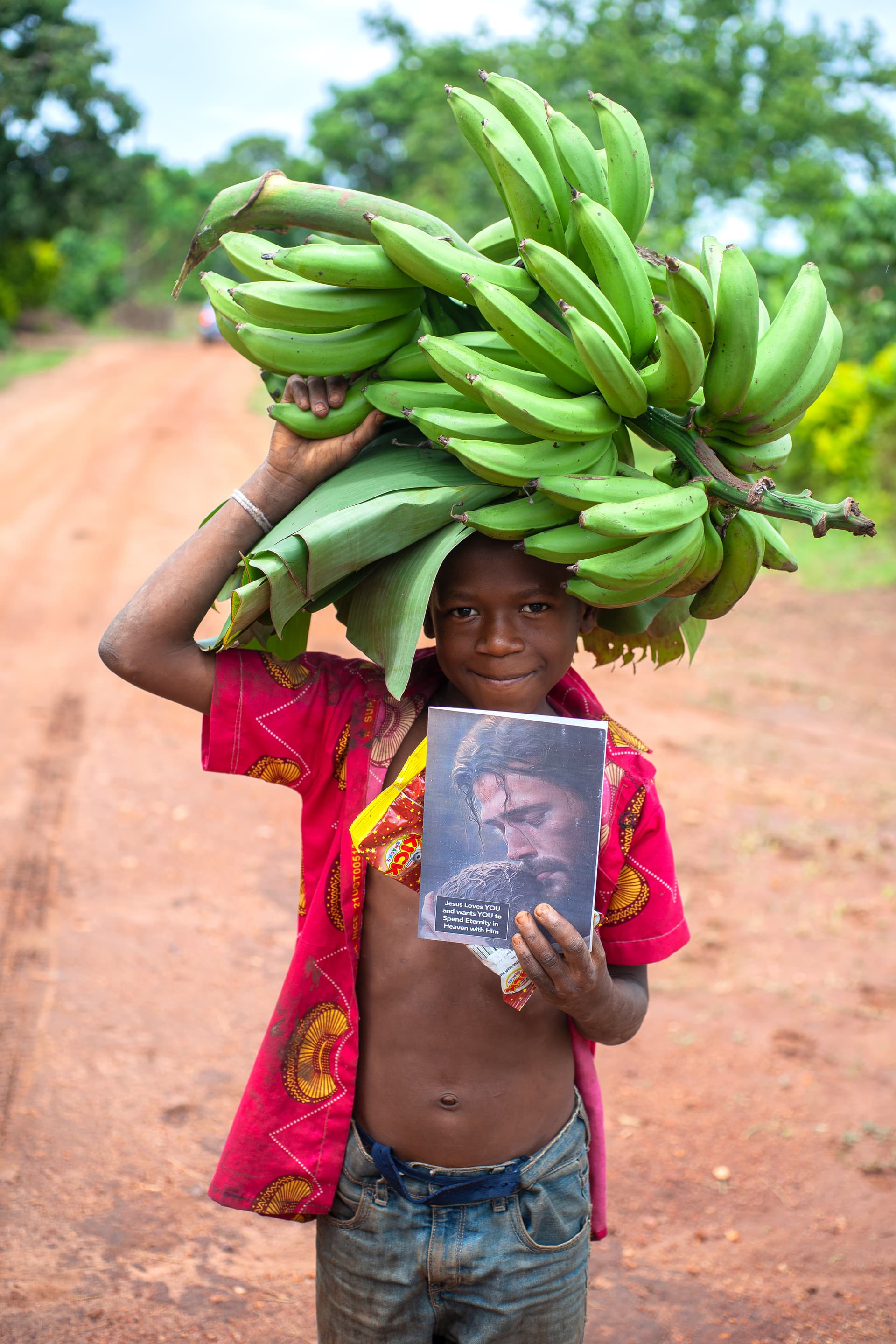 A boy carries a large bunch of green bananas on his head while holding a photo card.