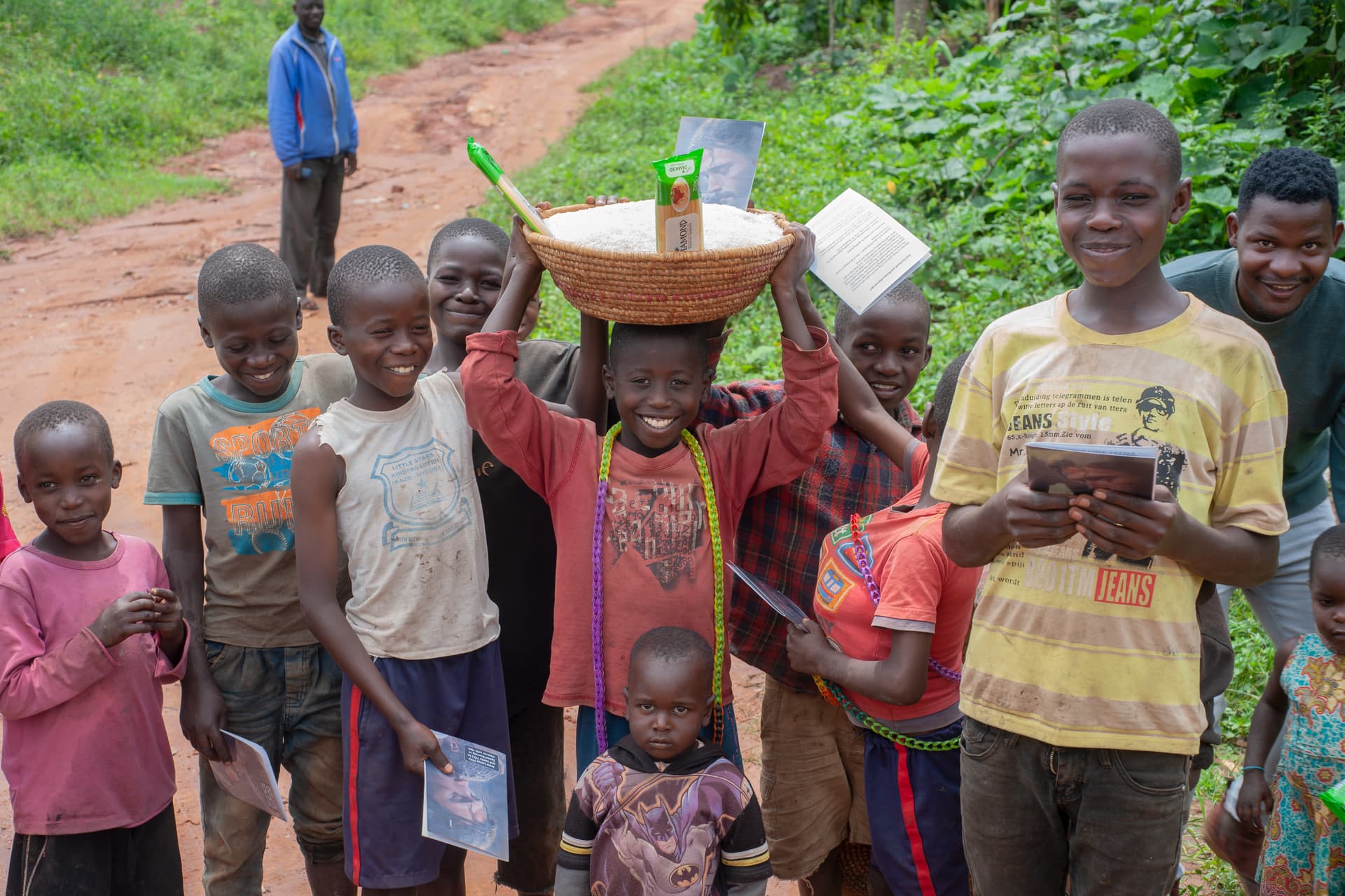 A group of children and a woman gather on a dirt road, one carrying a basket of food overhead.