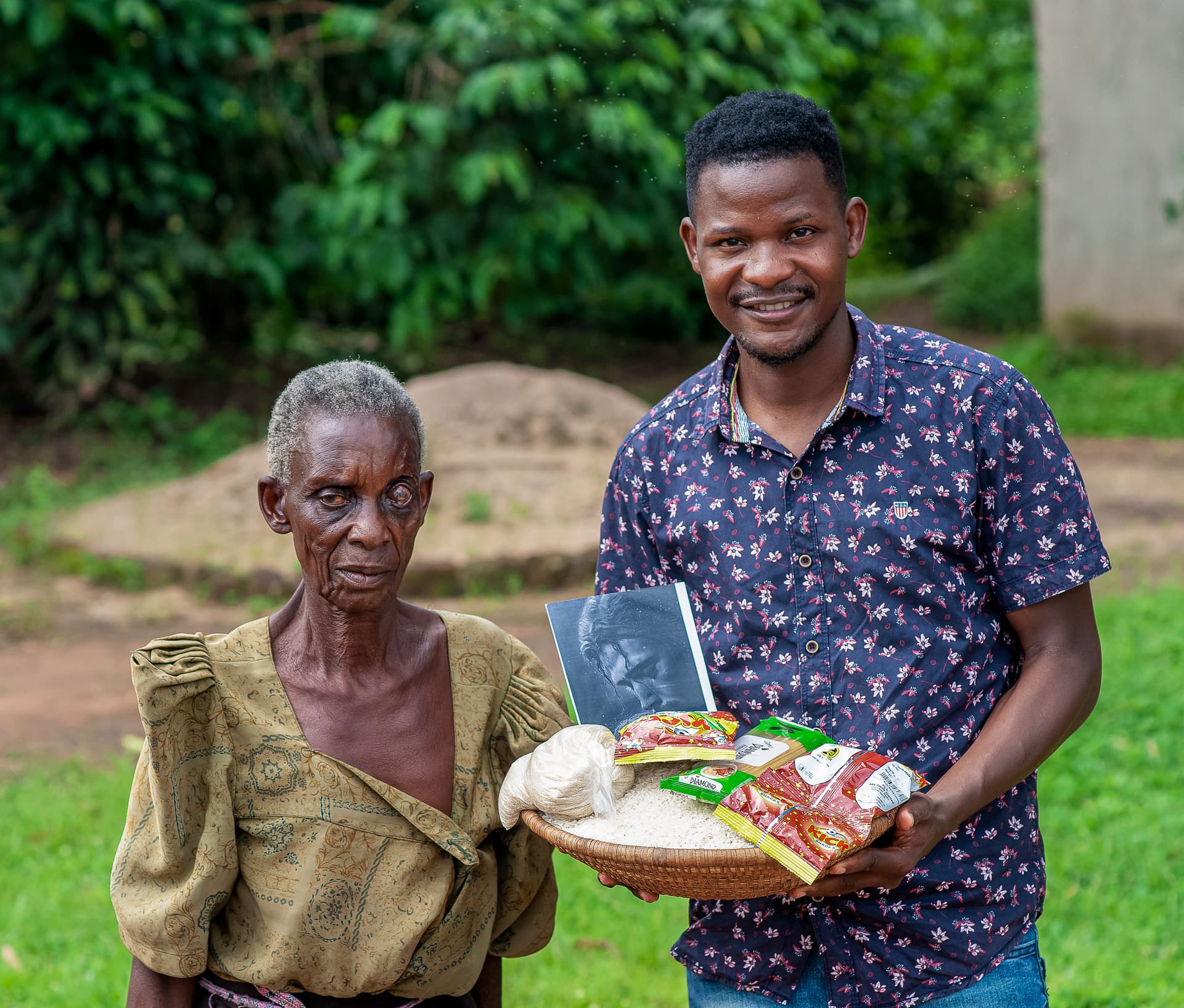 A man in a patterned shirt stands with an elderly woman, both holding a basket of food gifts, smiling.