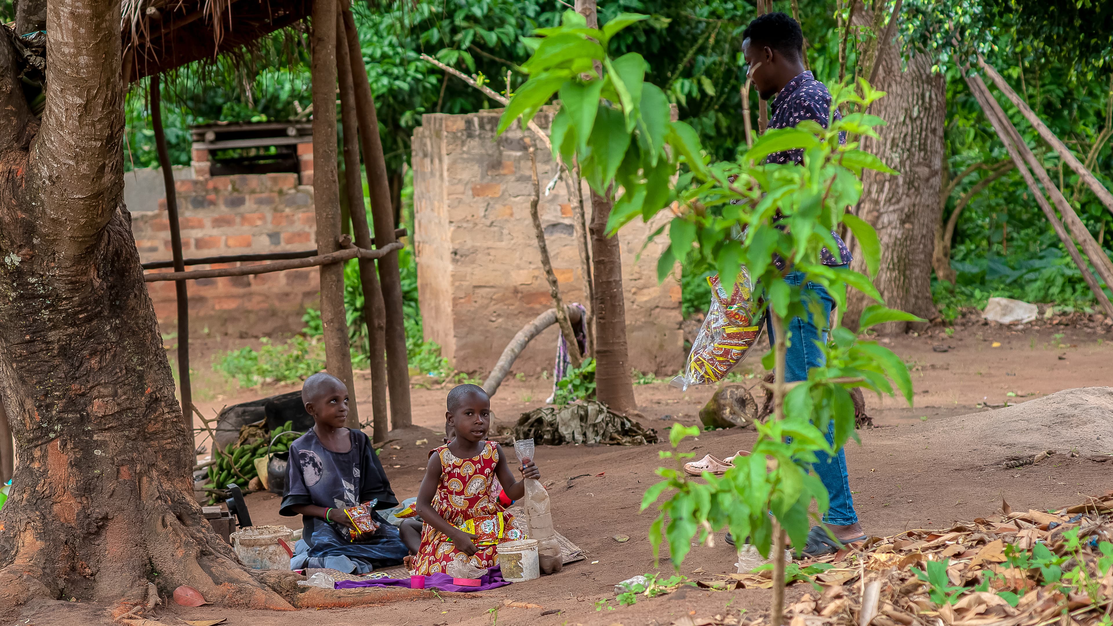 Children sit together under a thatched shelter in a village compound.
