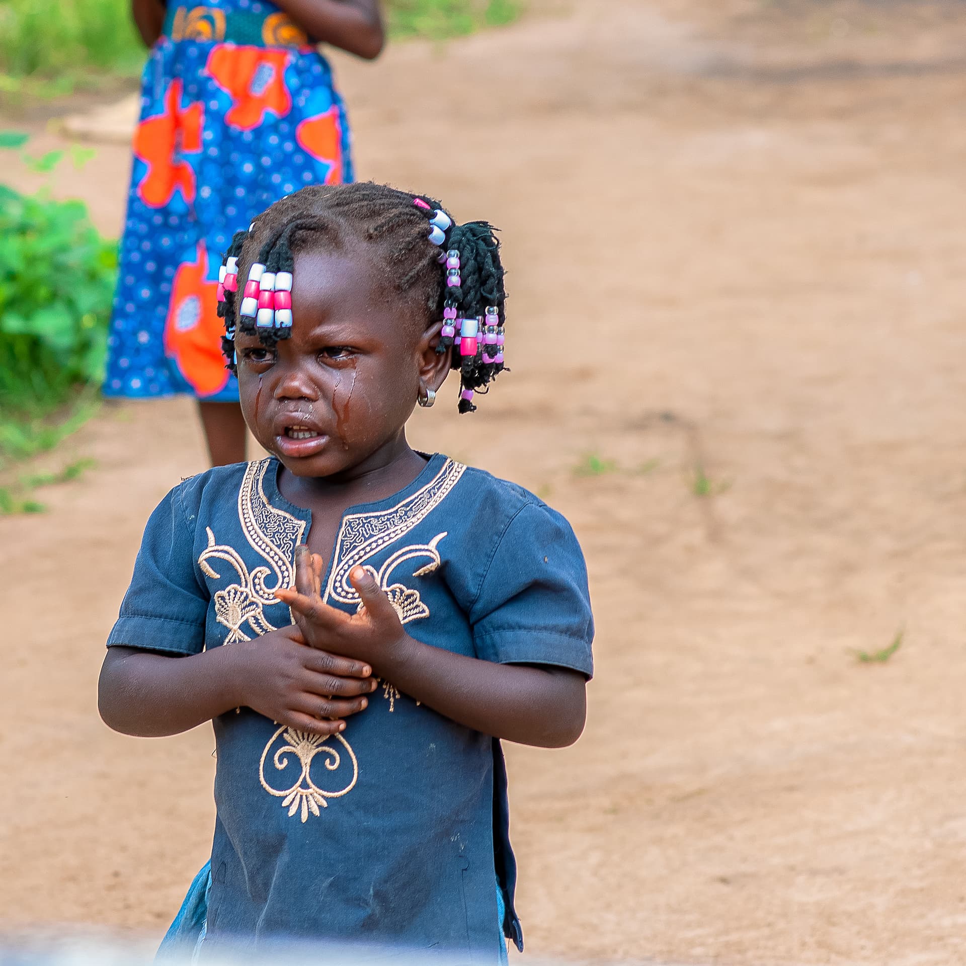 A young girl with hair clips looks toward the camera near a village home.
