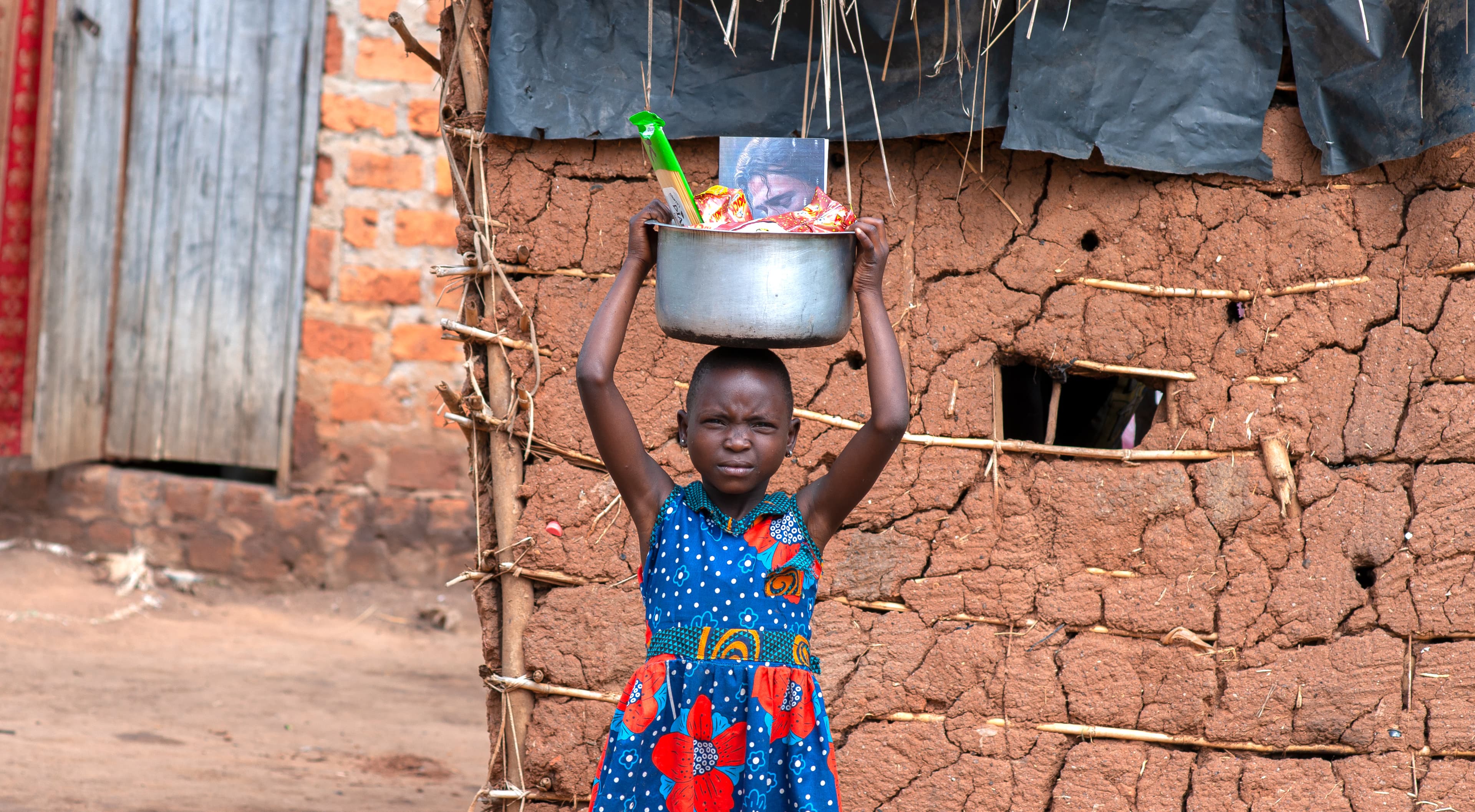 A child carries a metal pot on their head next to a brick wall.