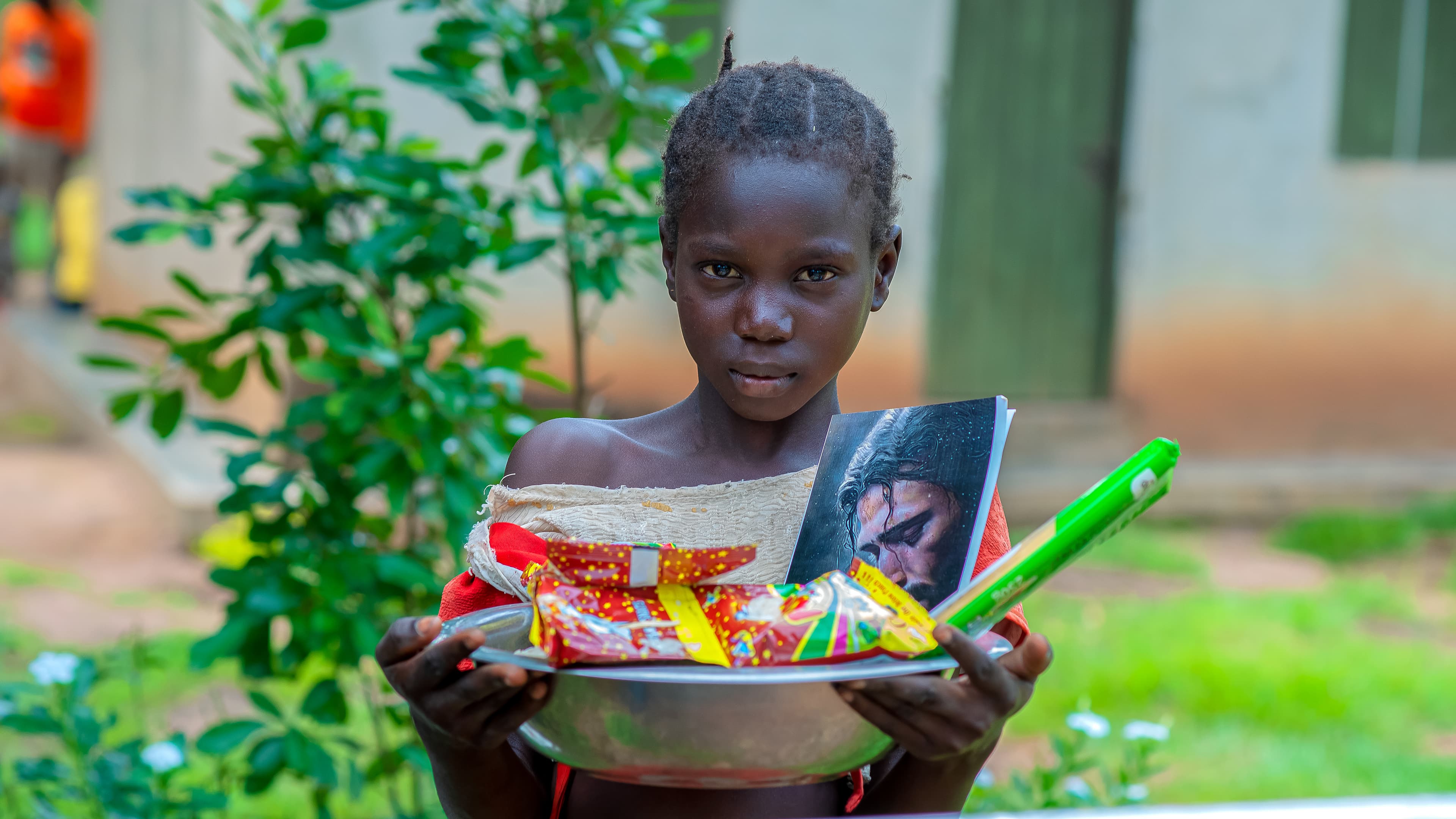 A girl holds a bowl with food items and a small photo, standing outside a building.