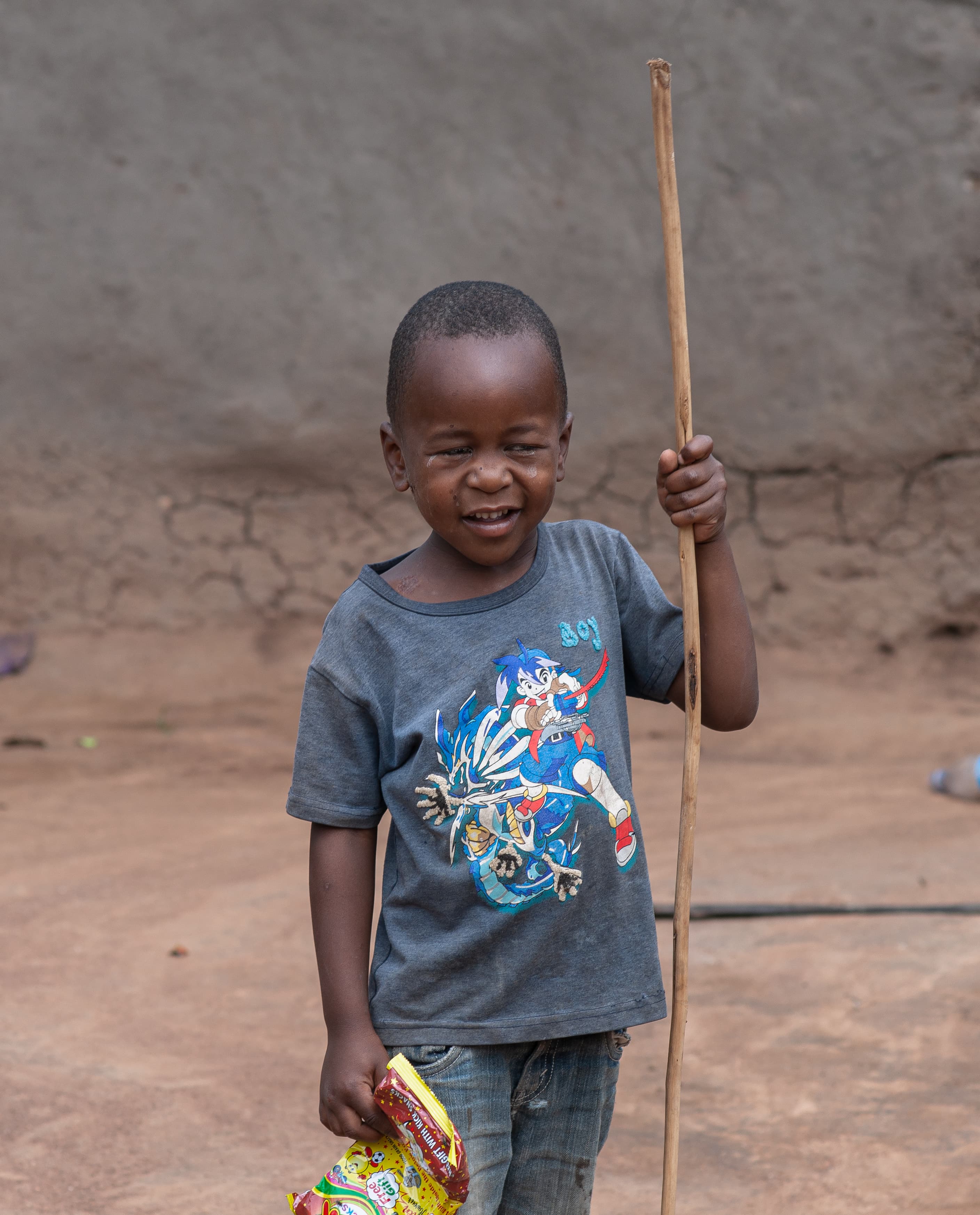 A boy holding a walking stick stands outside a mud house with a food gift bag.