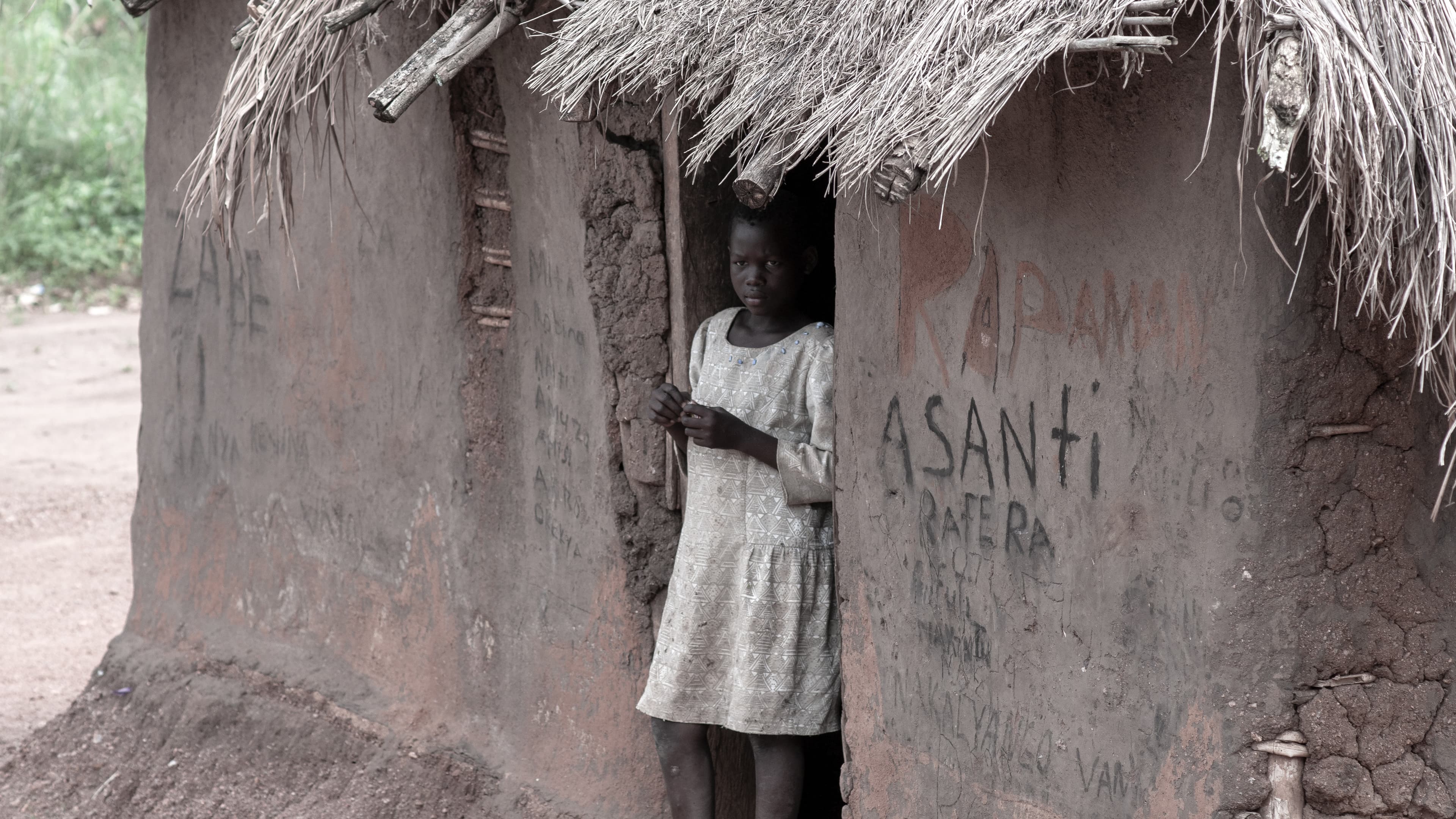 A girl stands alone in a mud hut doorway with the word ASANTI painted on the wall.