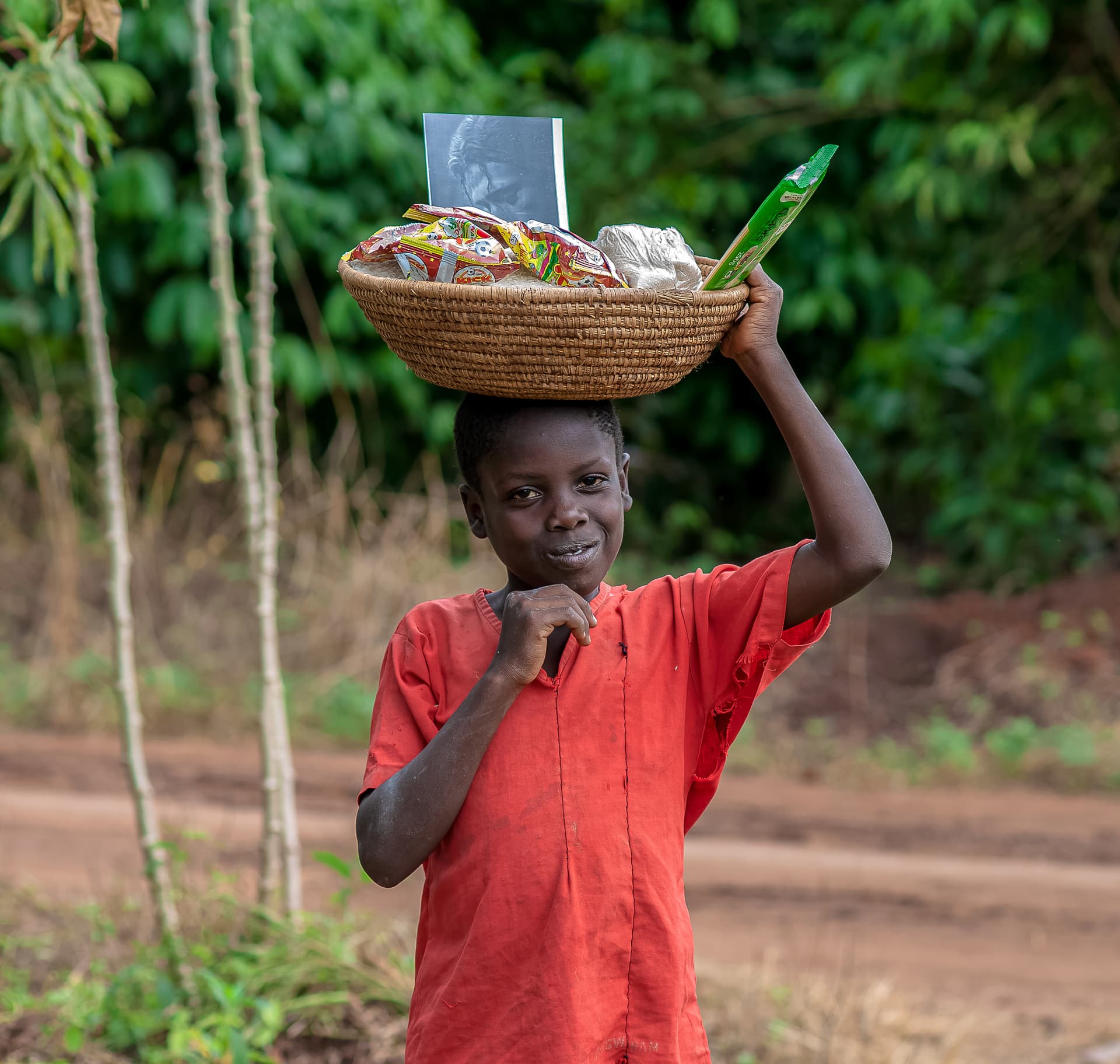 A boy holds a basket of food gift items above his head while standing on a path.