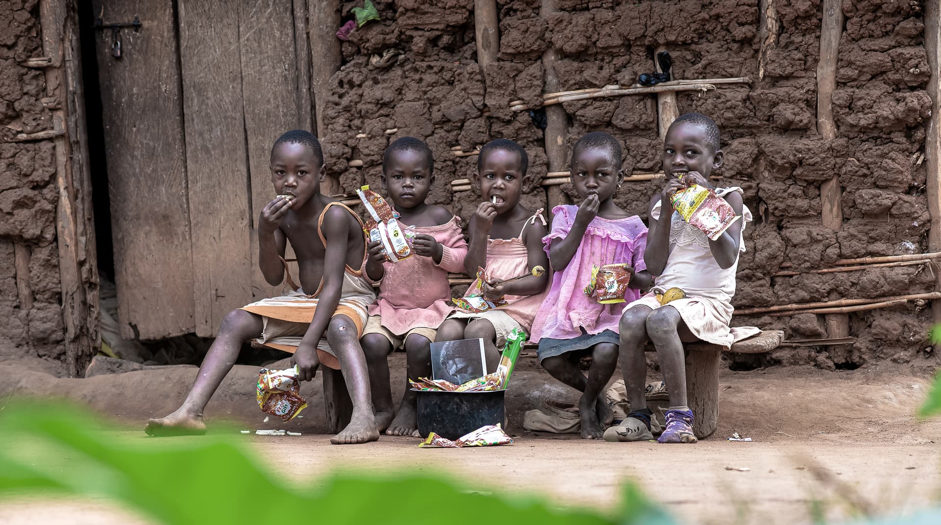 A group of children sit together on a bench outside a mud-walled home.