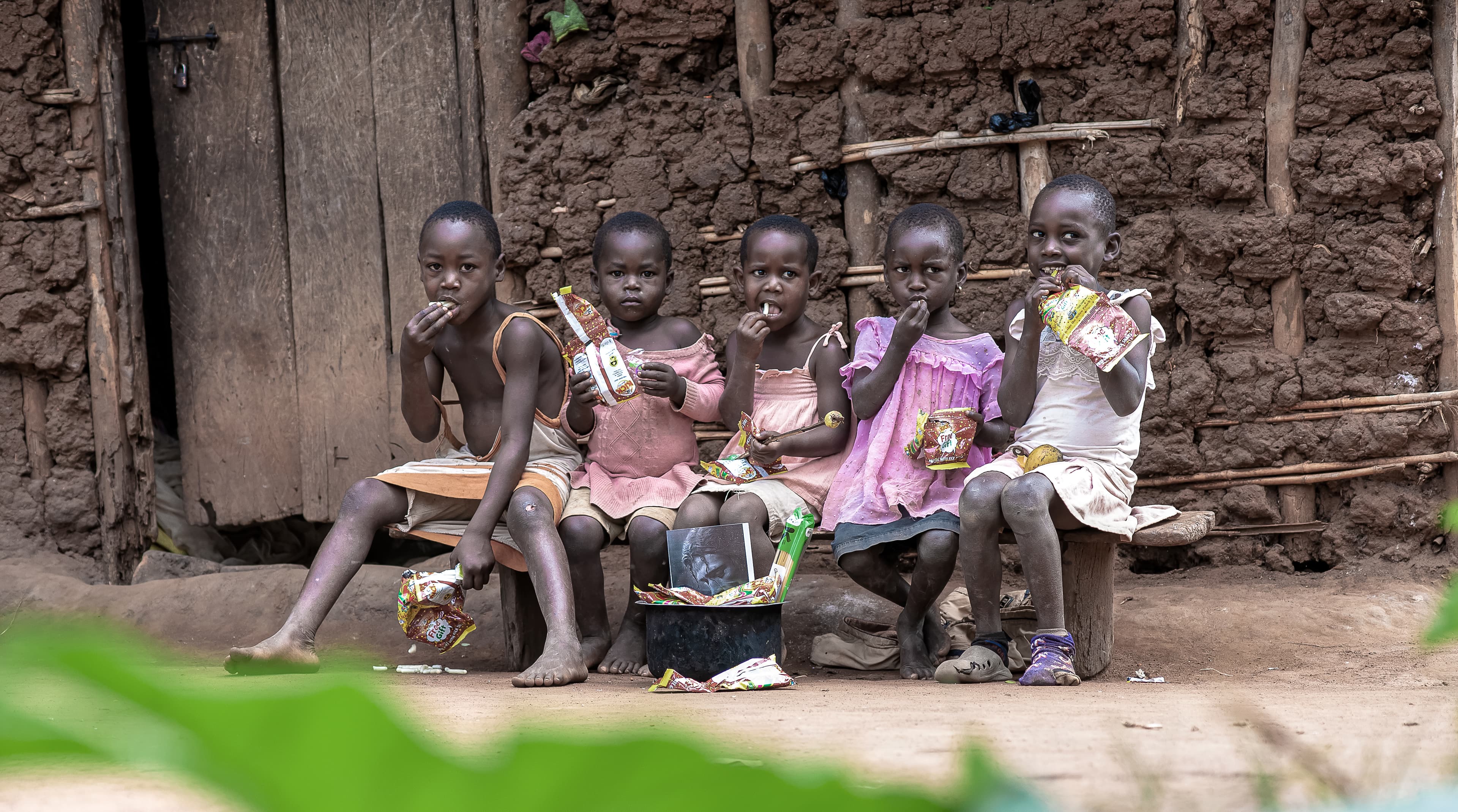 A group of children sit together on a bench outside a mud-walled home.