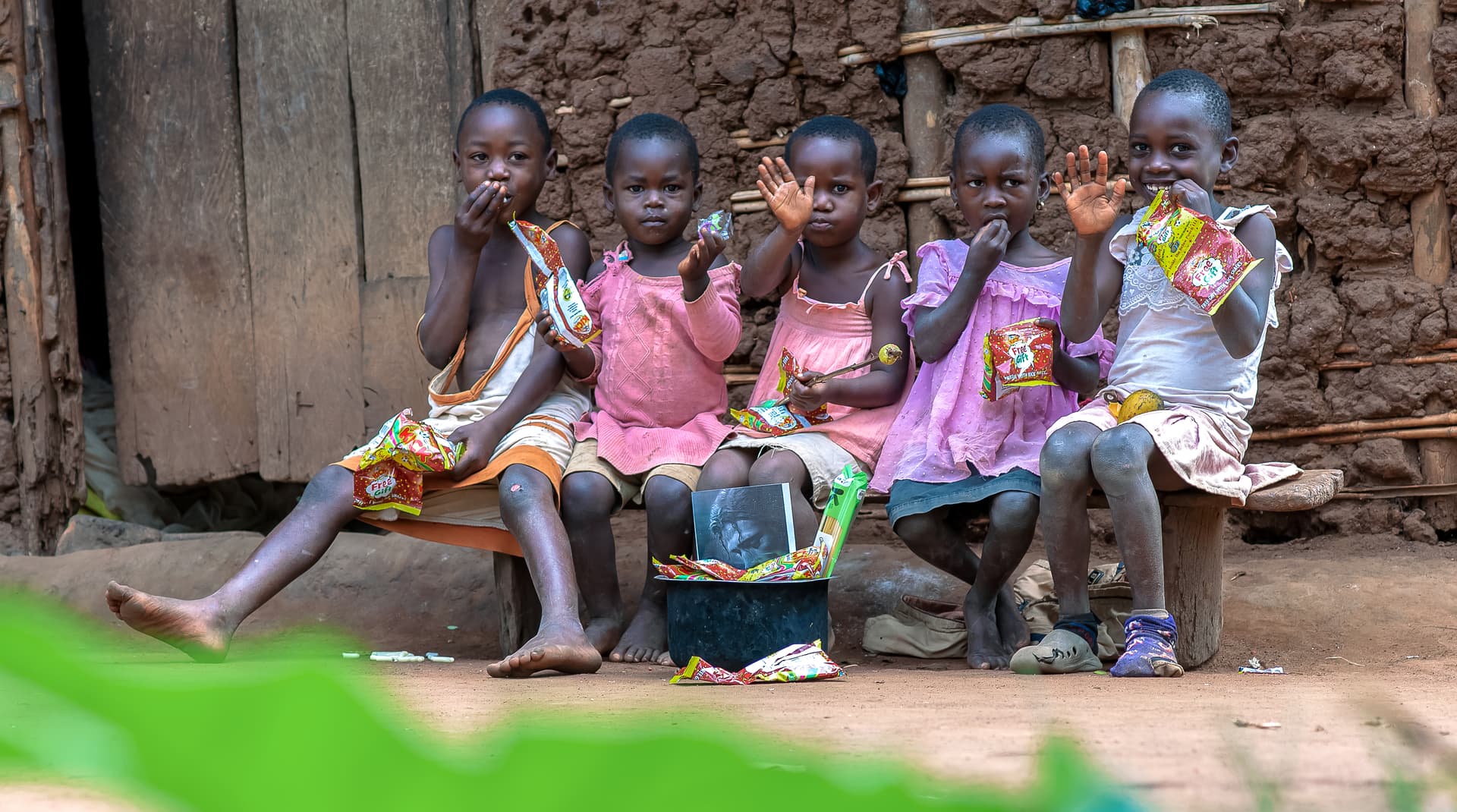 A row of children sit against a mud-brick wall outdoors, wearing colorful clothing.