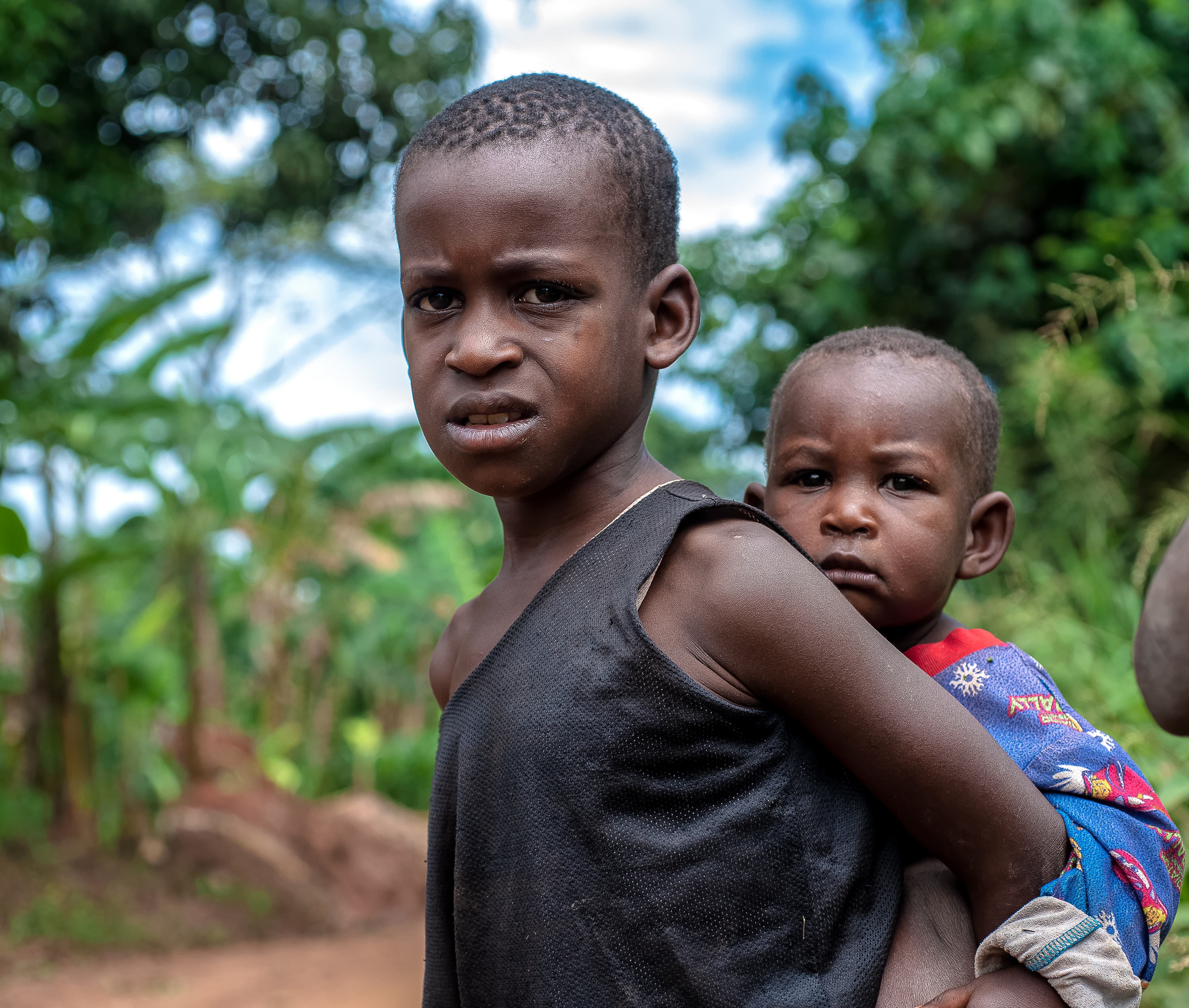 An older child carries a younger child on her back along a dirt path.