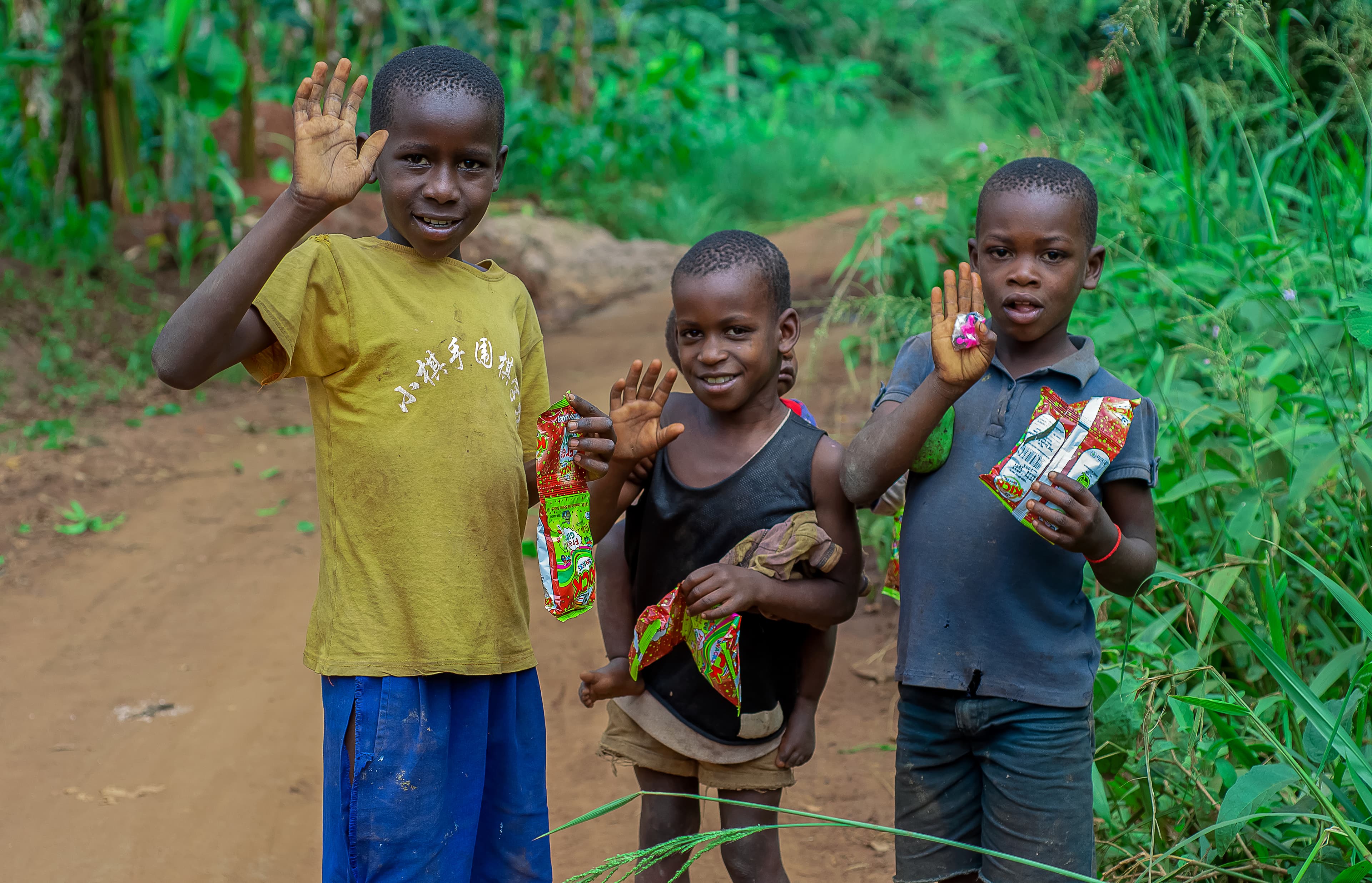 Three children stand on a dirt path, one waving, holding food gift items.