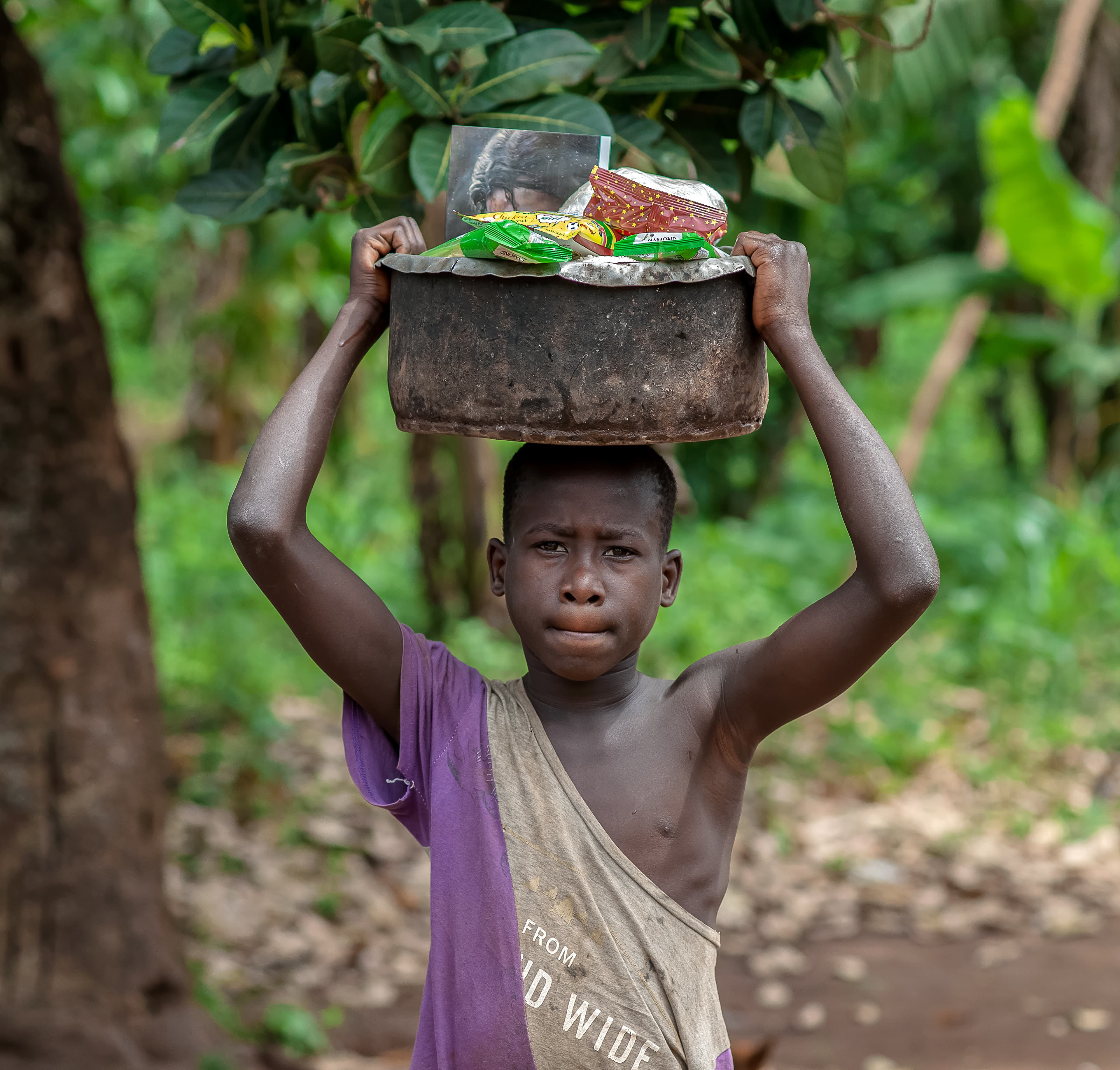 A boy carries a pot of food gift items on his head outside a brick building.