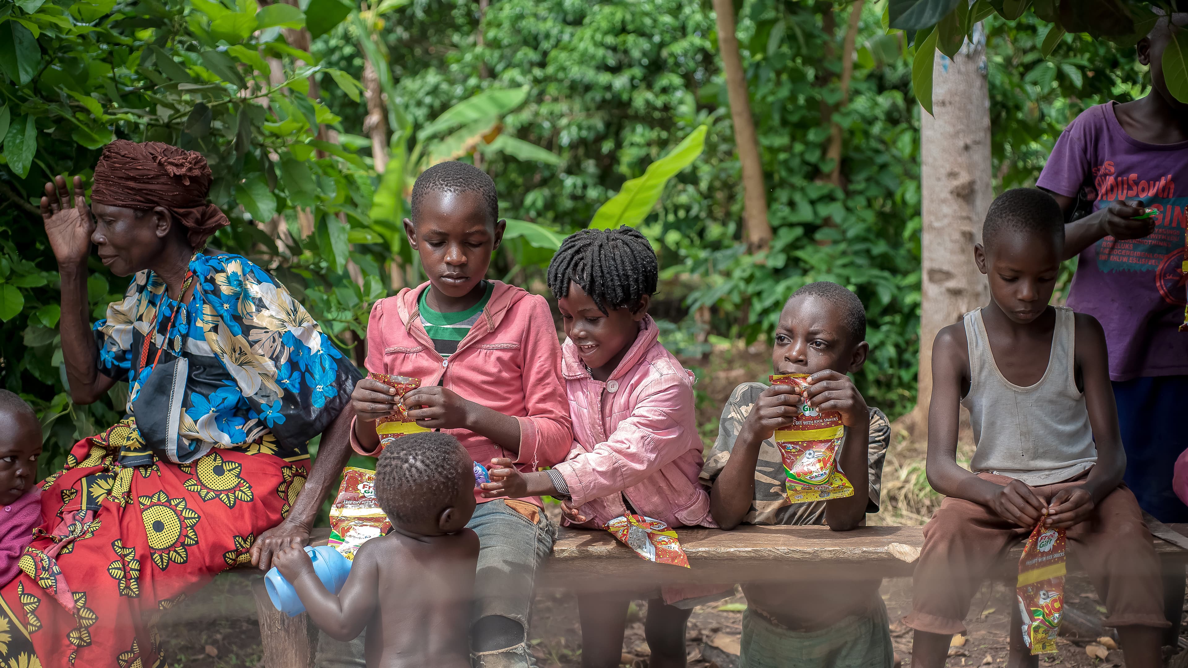 Woman in a colorful dress sitting with children eating from bowls outdoors