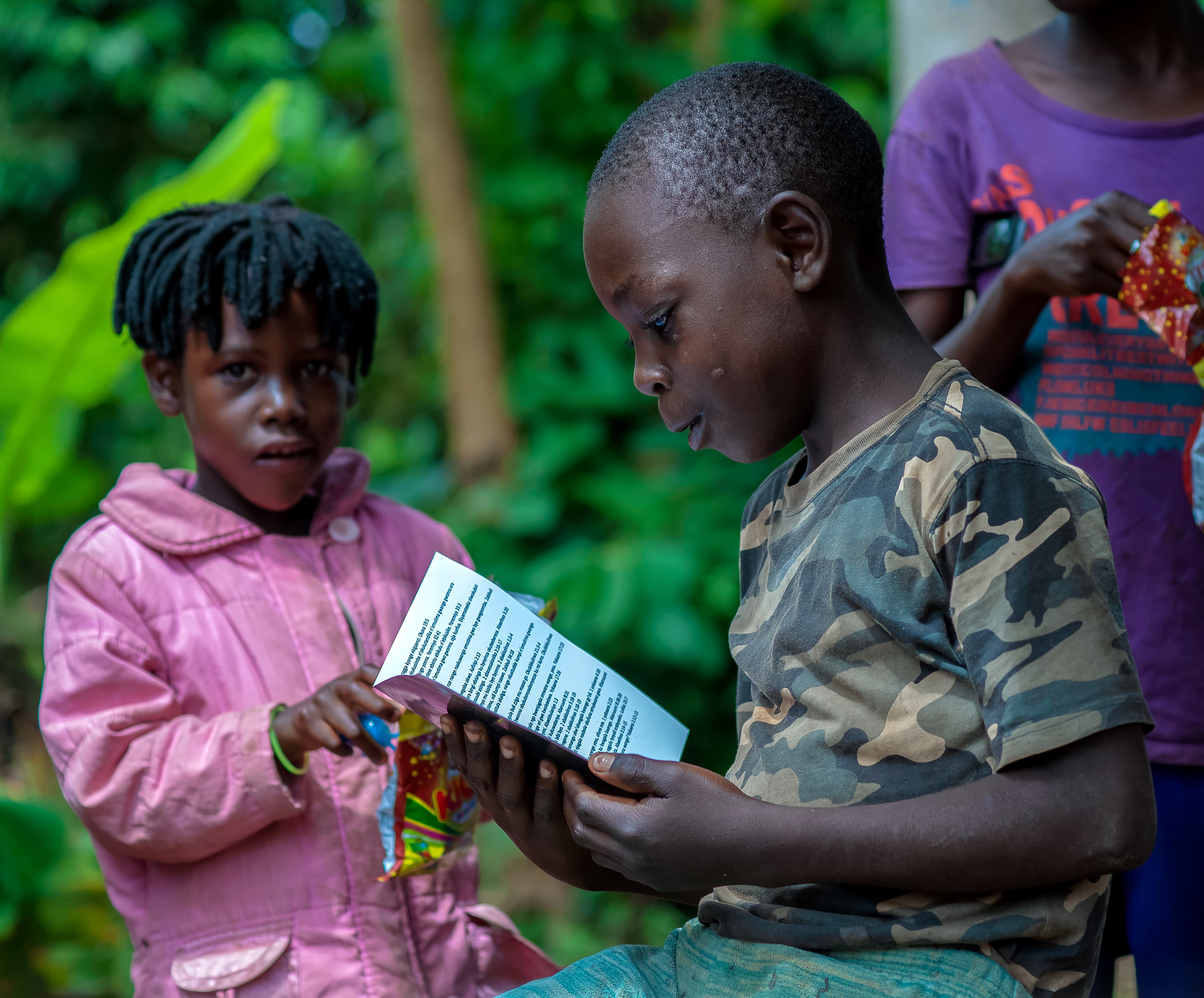 Two girls stand together in greenery, one reading from a printed photo booklet.