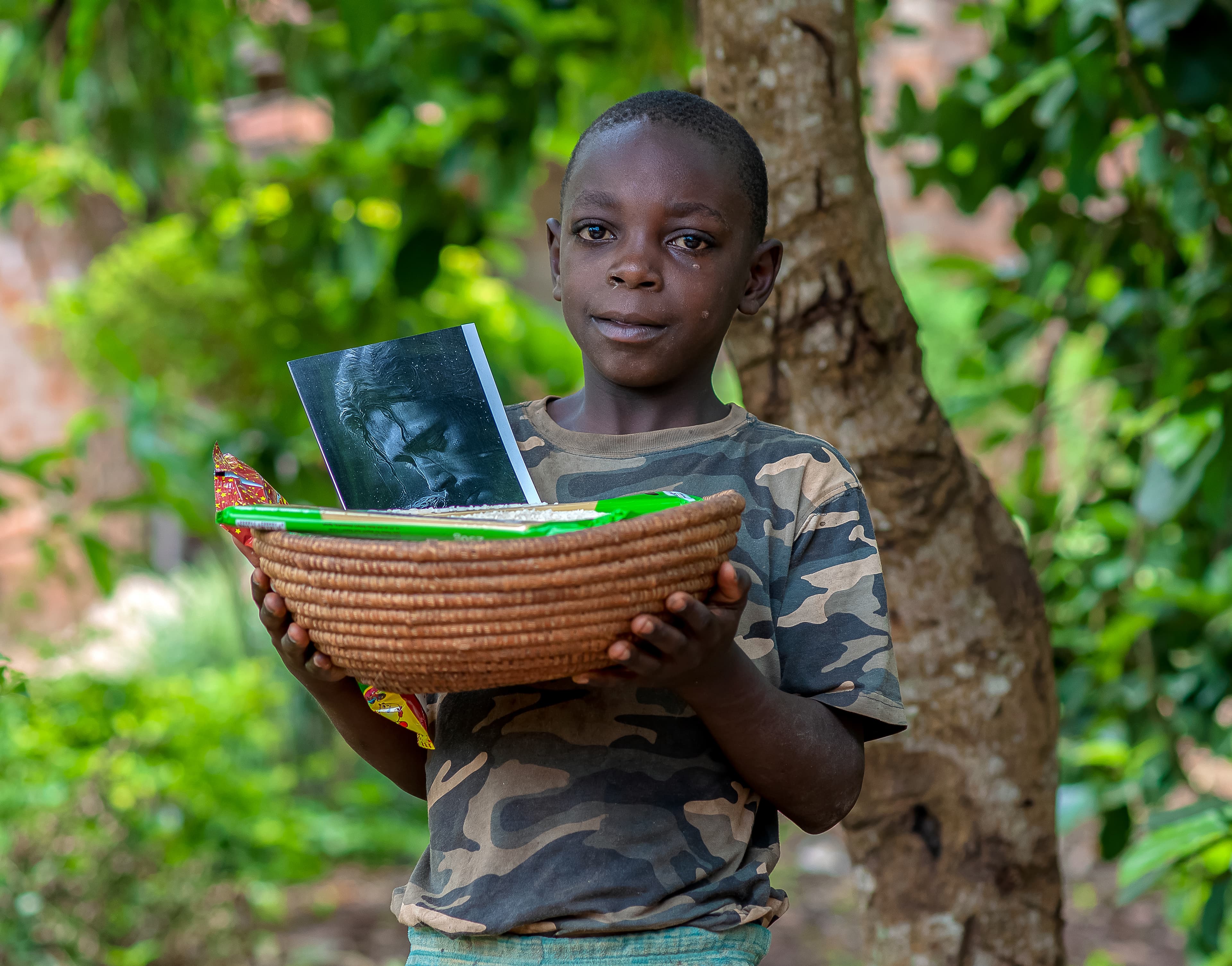 A boy holds a basket of food gift items with a photo, standing beside a tree.