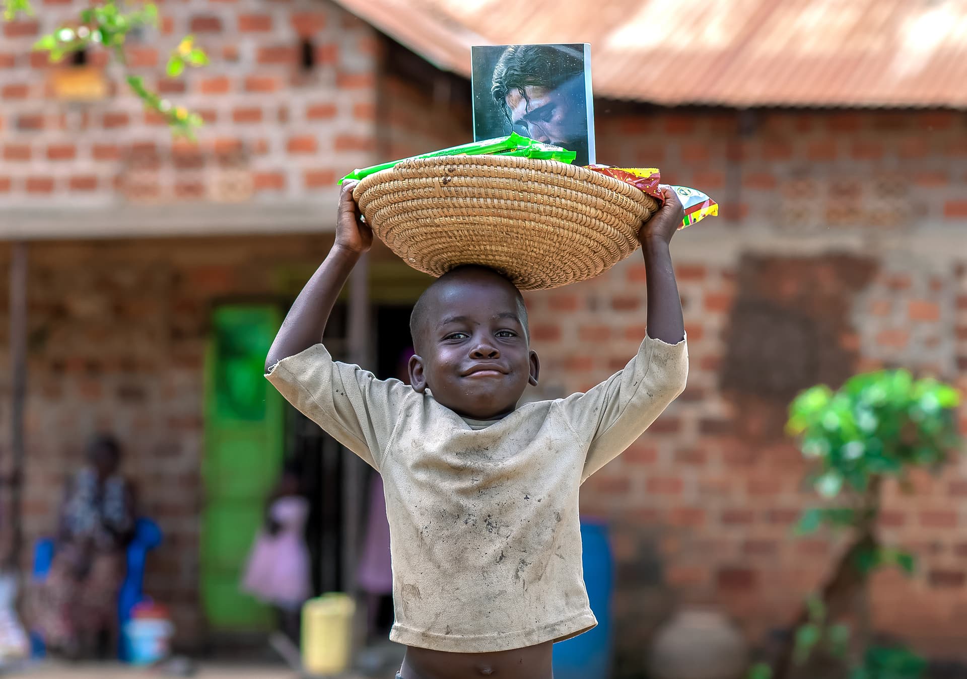 A boy holds a basket with food items and a printed photo above his head near a brick building.