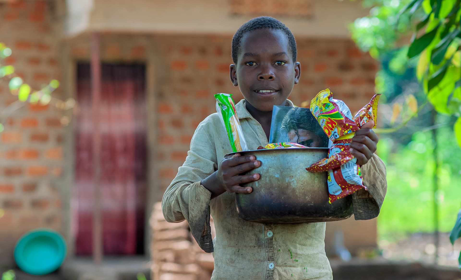 A boy stands near a brick house holding a metal pot and food gift items.