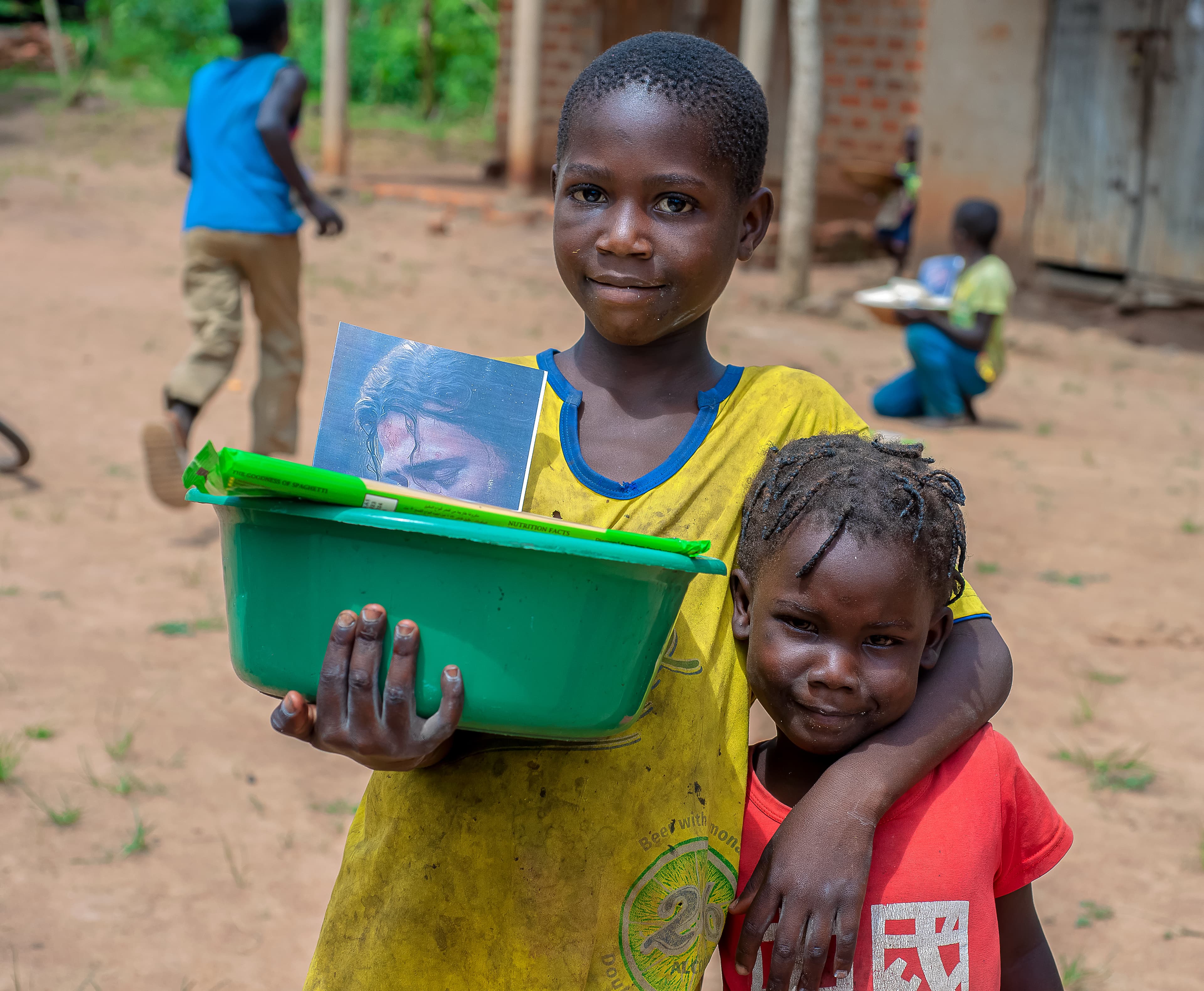 A girl holding a green basin hugs a smaller child in red on a dirt path.