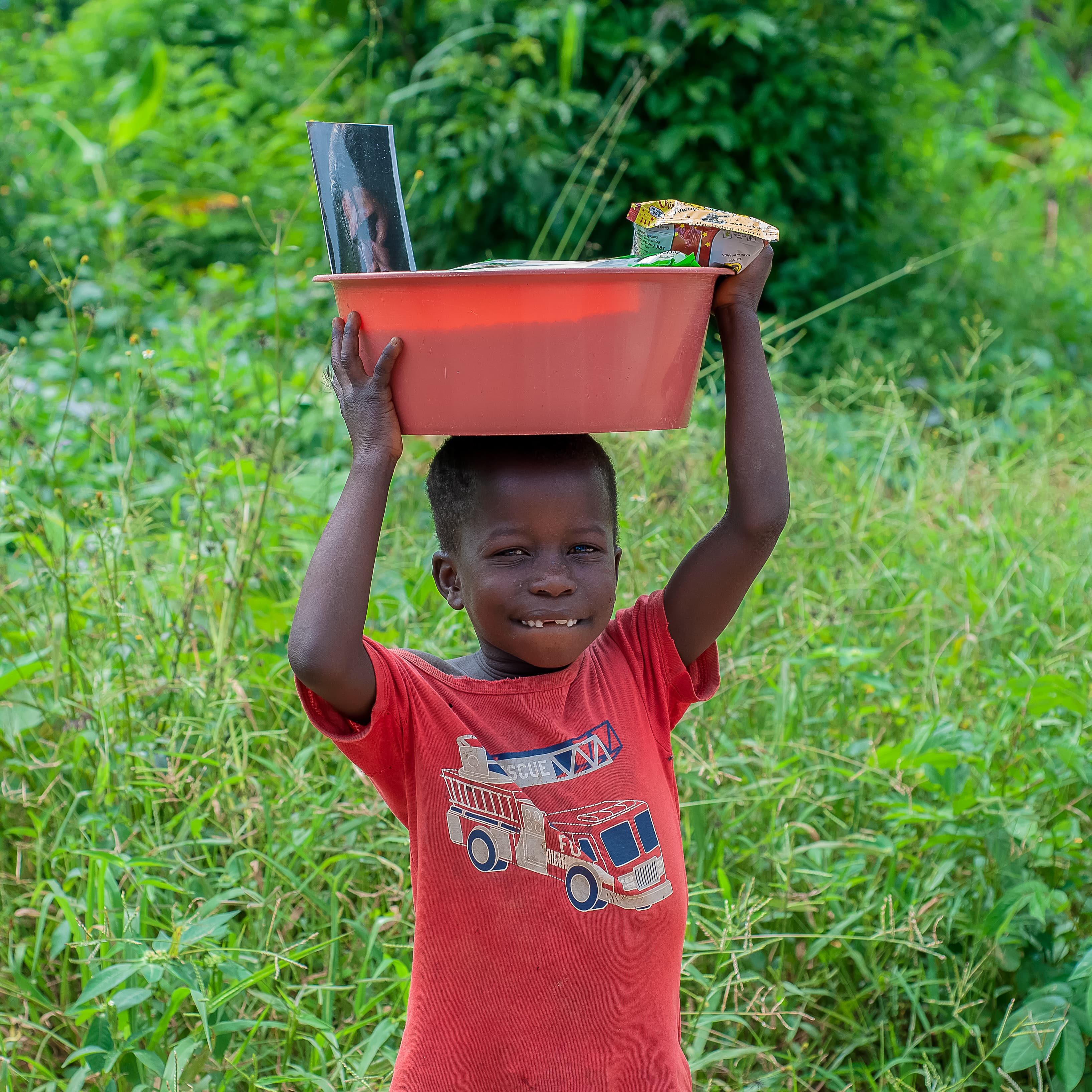 A boy lifts an orange plastic basin above his head while standing in tall grass.