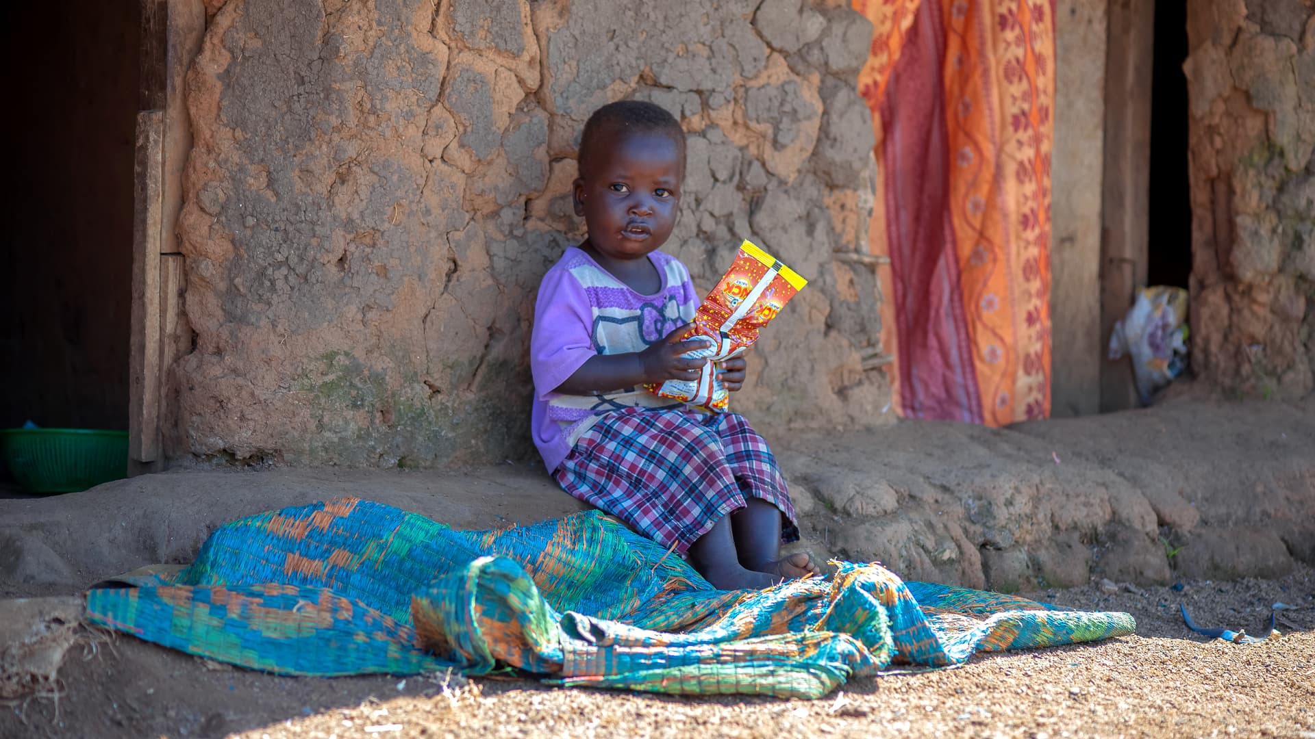 A child sits on a colorful mat outdoors holding a food item.