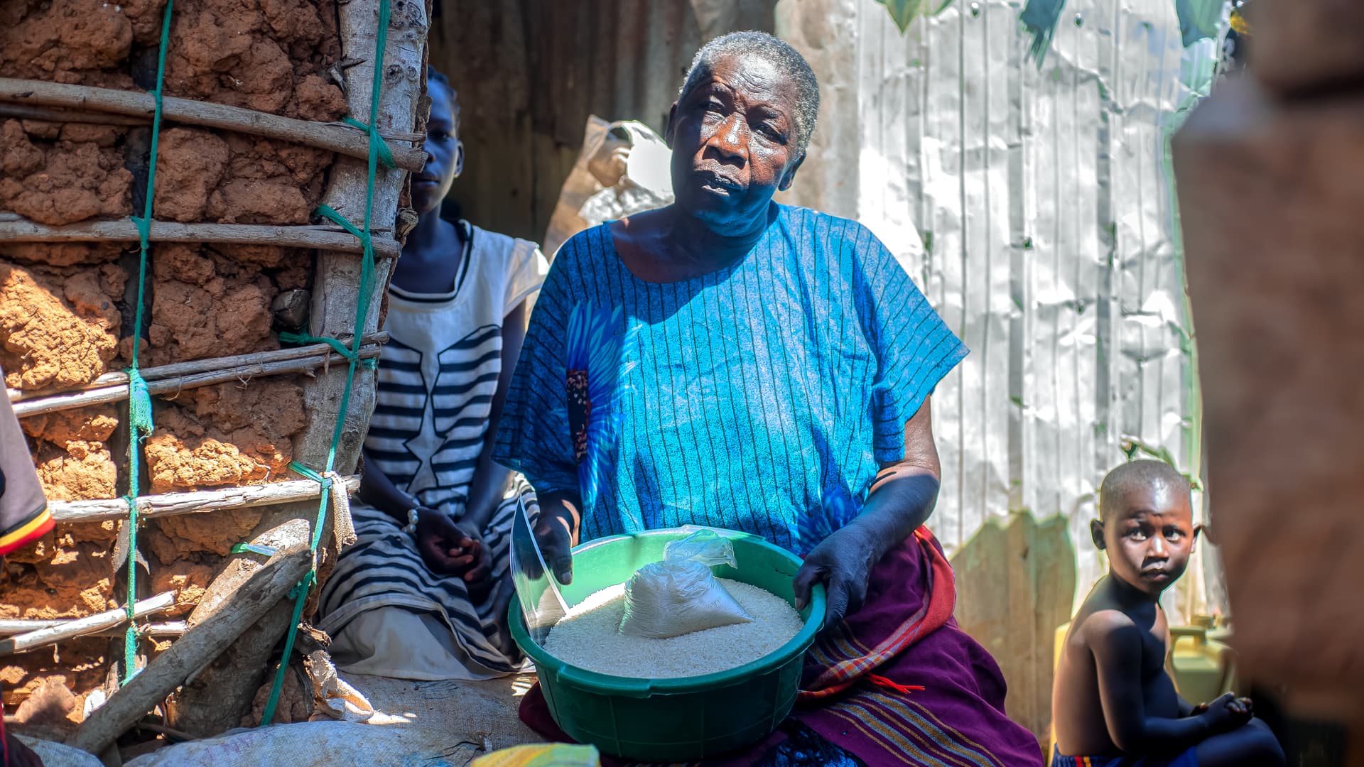 An elderly woman sits outdoors holding a green pot, with a child nearby.