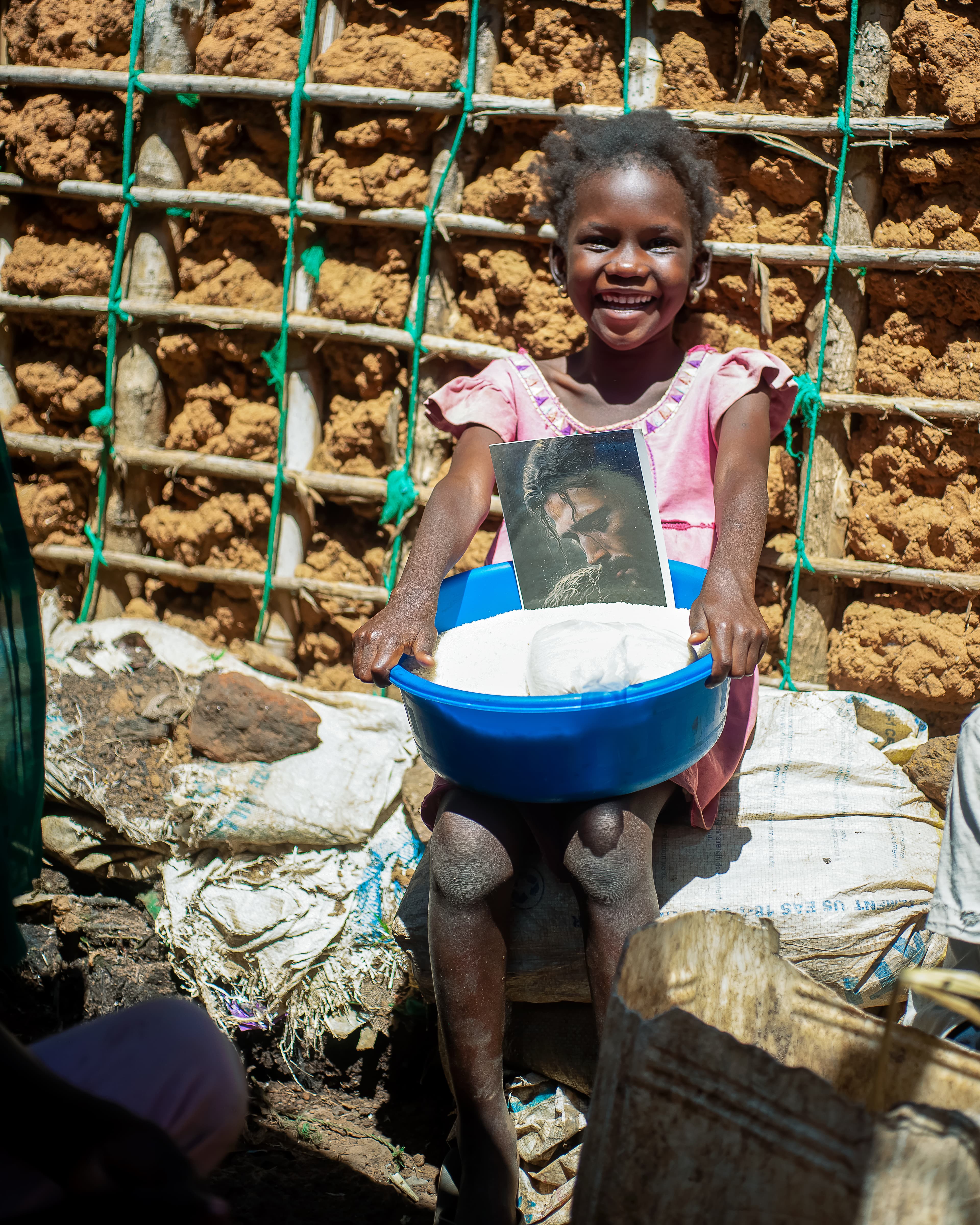 A girl sits near a wooden frame holding a basin with food items and a photo.