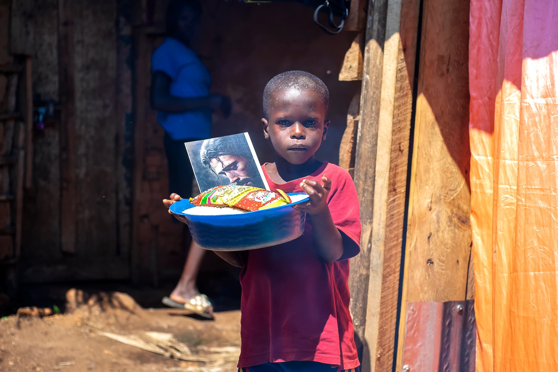 A boy in a red shirt stands in a curtained doorway holding a basin of food gifts.