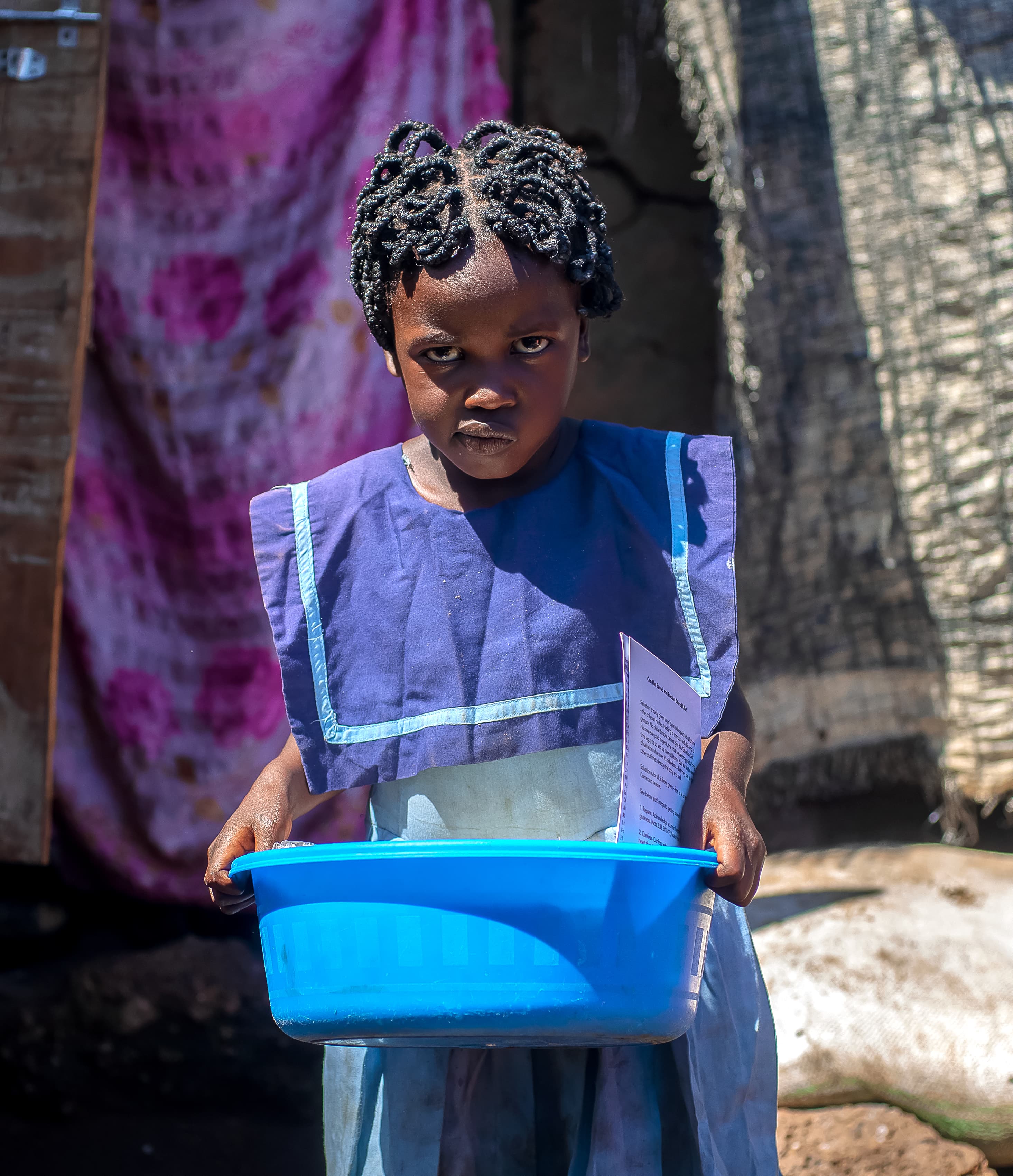 A girl in a patterned dress holds a blue basin outdoors.