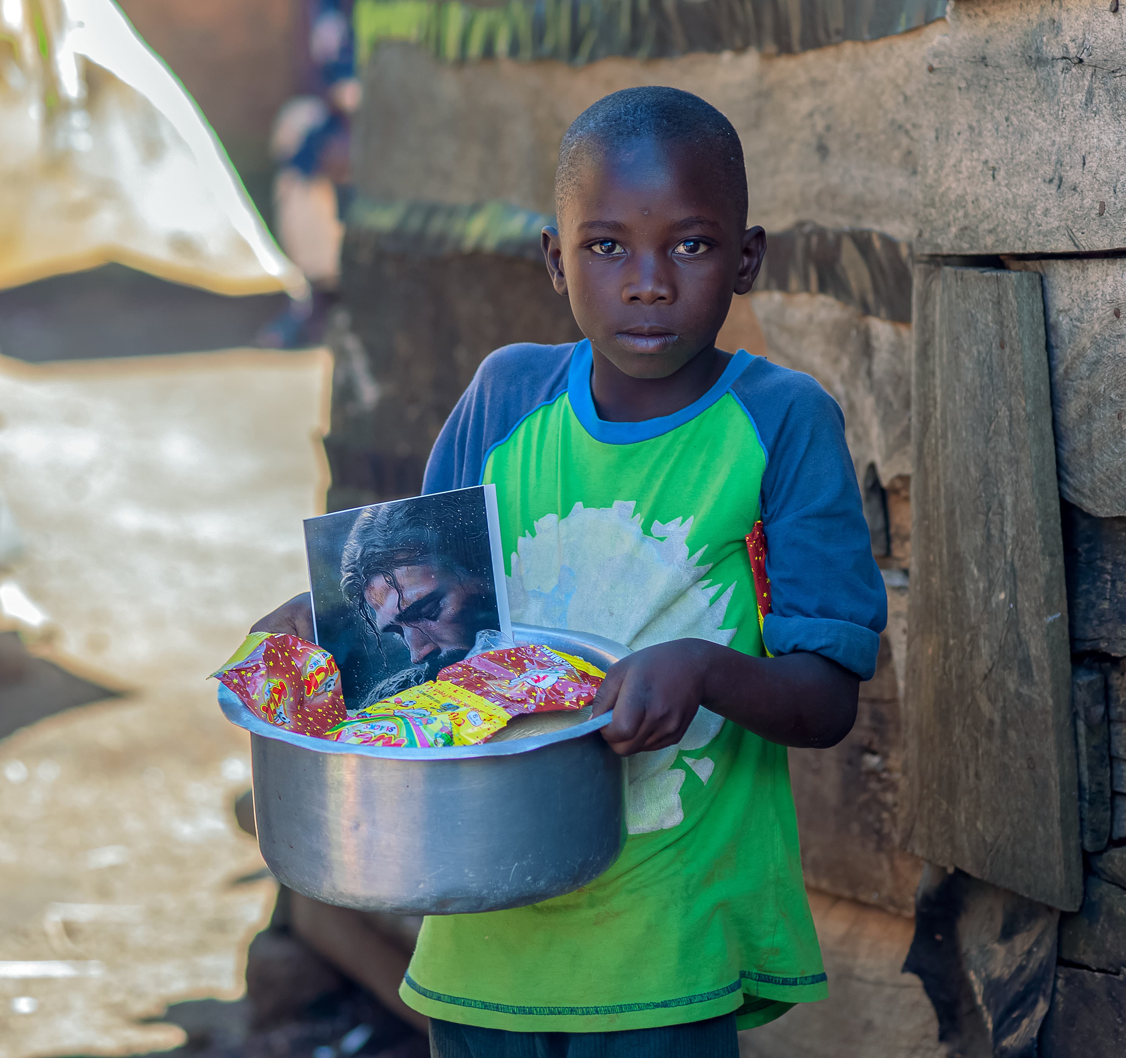 A boy in a green shirt holds a metal basin of food gifts outside a shed.