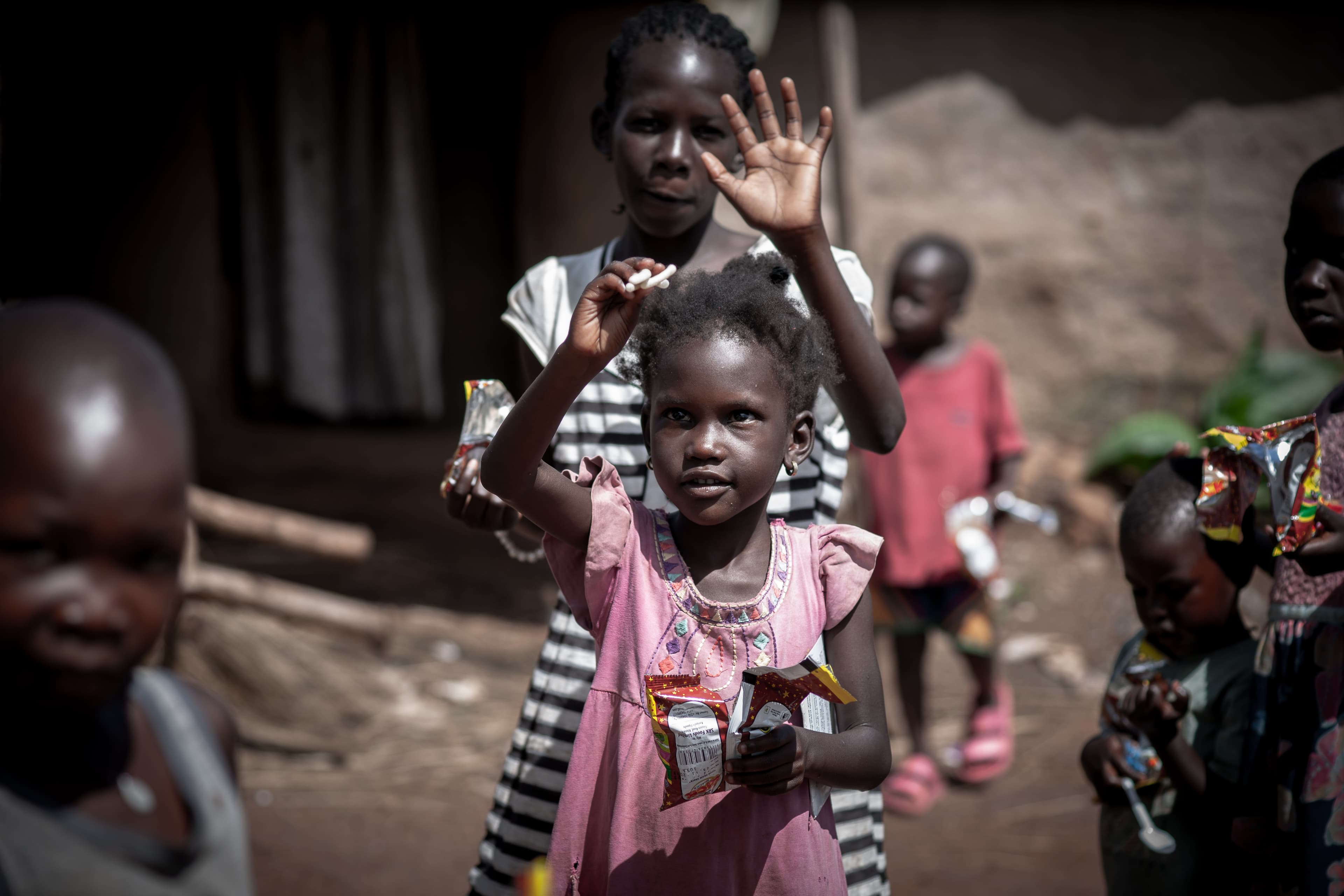 A group of children stand outside a brick house, one of them waving.