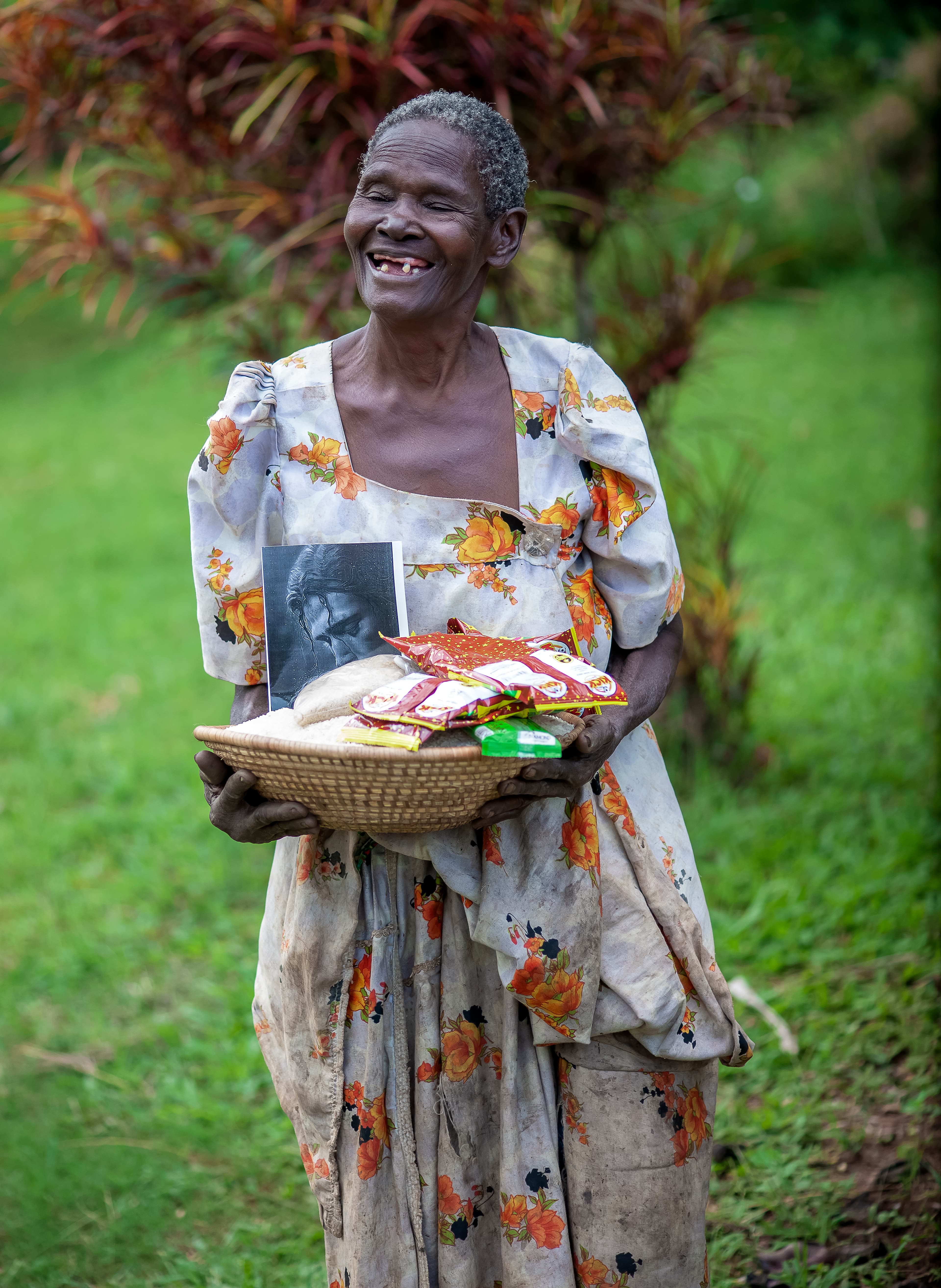 An elderly woman smiles while holding a basket with food items and a small photo.