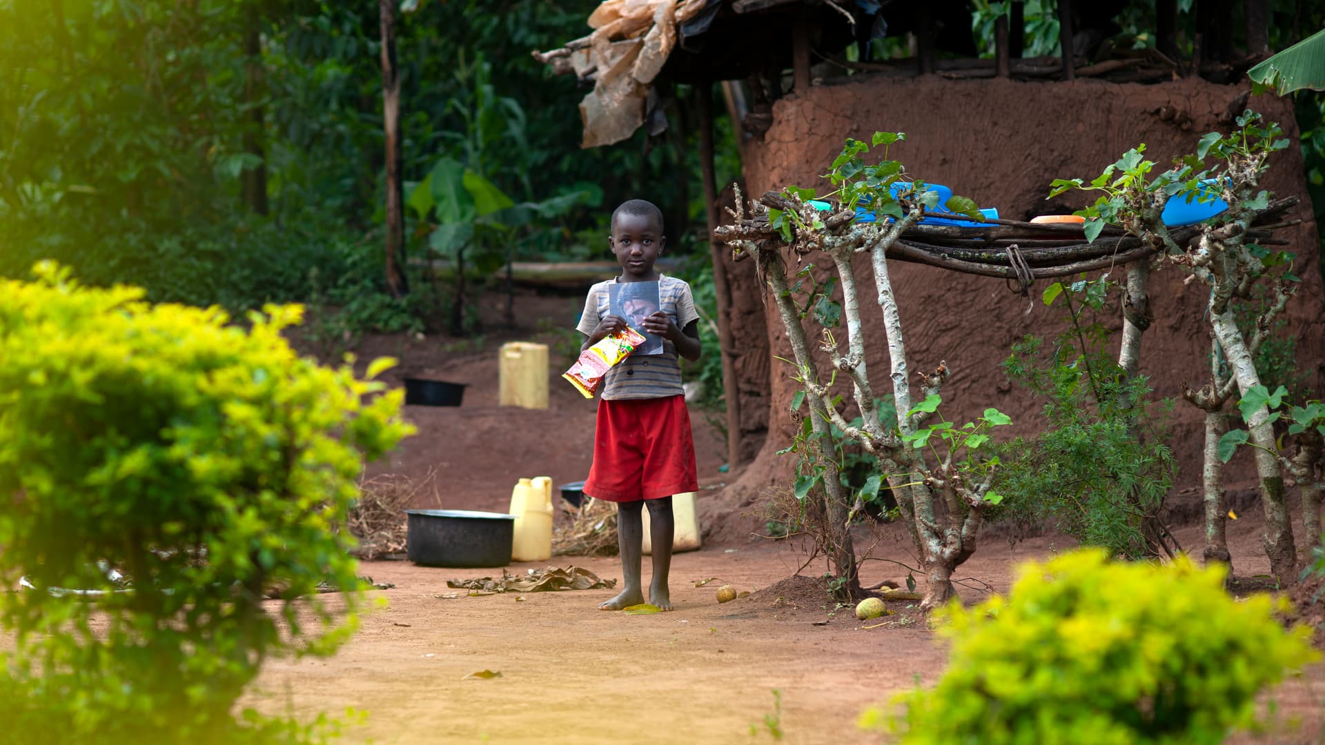 A child stands outside a mud and thatch house at dusk, seen from a distance.